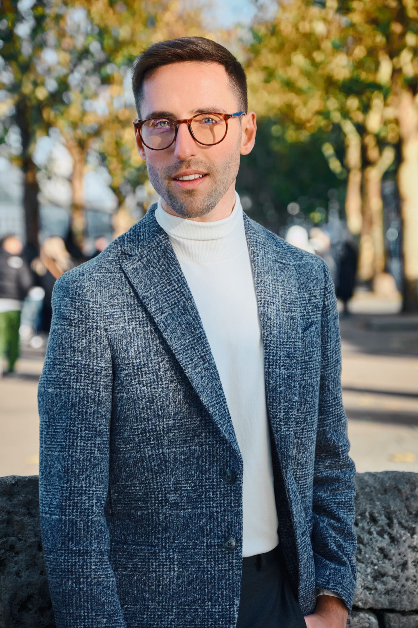 A man in a blue blazer and white turtleneck stands in front of a stone wall, smiling and looking at the camera.
