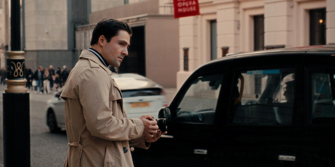 Actor Robert Clooney, dressed in a beige trench coat and blue scarf, stands on a city street, holding a phone and gazing off to the side. A black car 