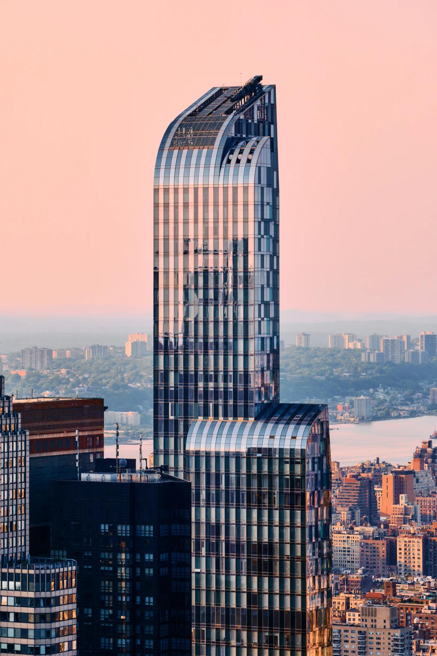A modern skyscraper with a curved glass facade and blue tinted windows dominates the cityscape, reflecting the pinkish-orange sky. The cityscape featu