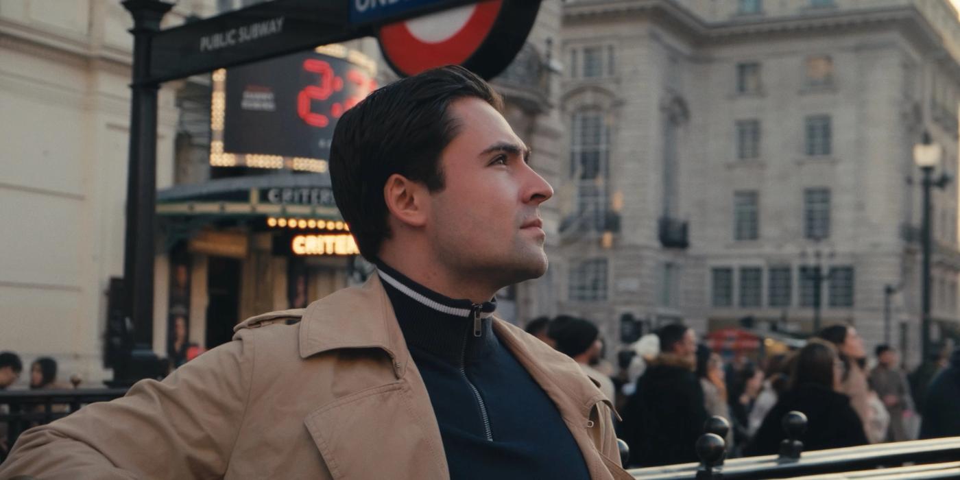 Man in beige coat, looking up at London Underground sign, surrounded by people and buildings.