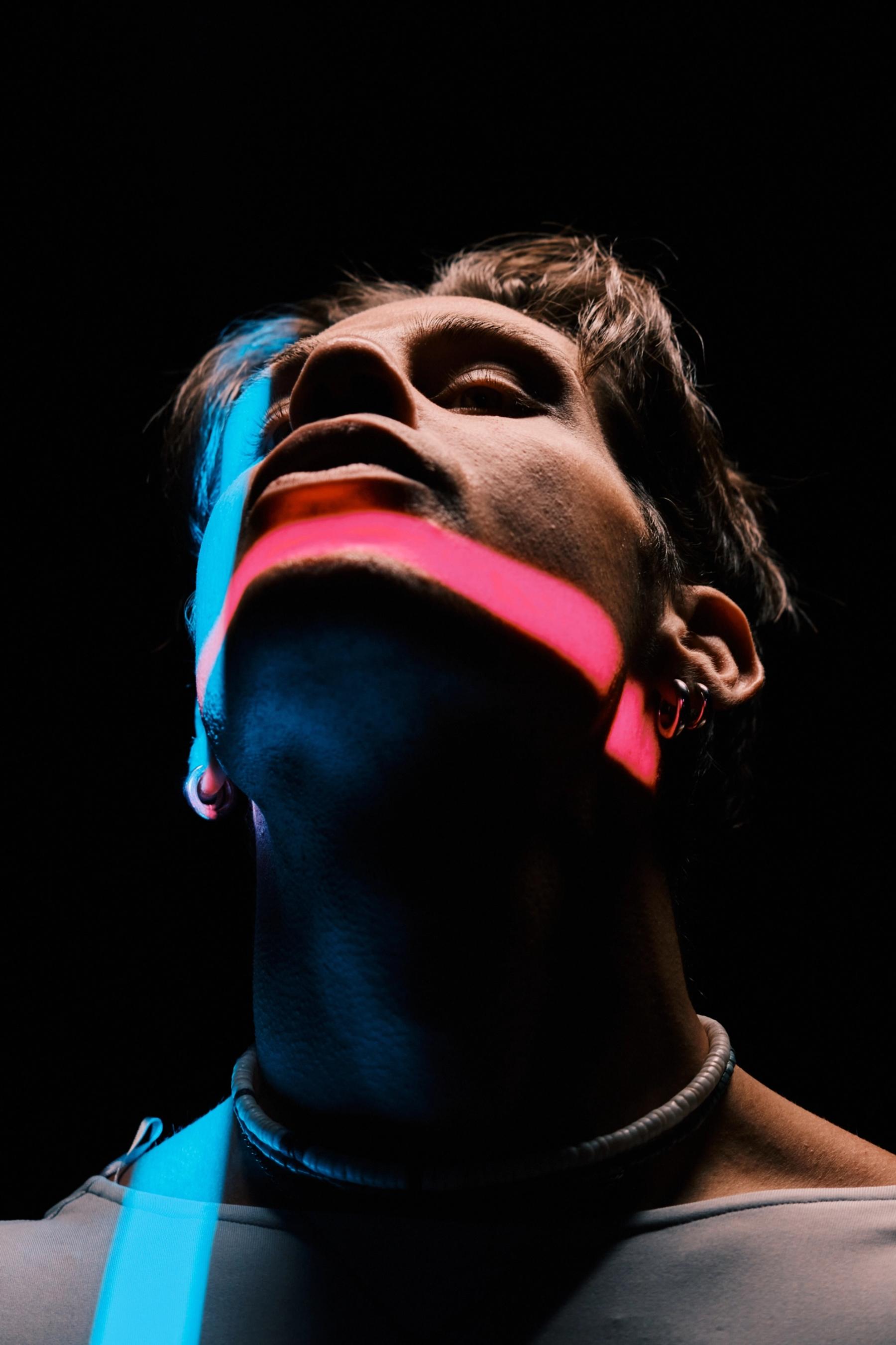 A man with a pink and blue neon-like collar, wearing a necklace, is looking upwards, with his eyes closed and mouth open, gazing at the sky.