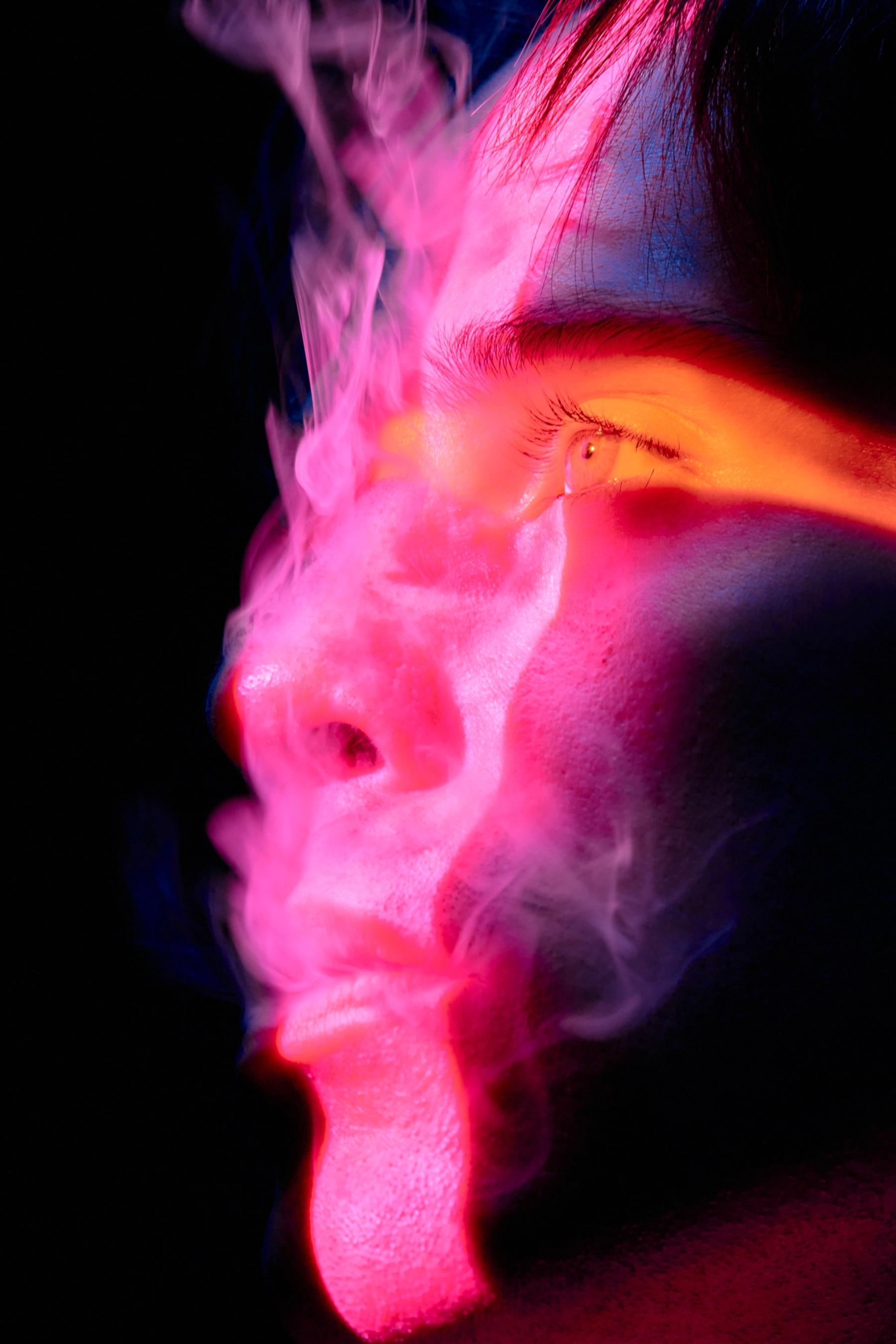 A close-up of a person's face, with pink and purple smoke emanating from their eyes, creating a mysterious and intriguing scene.