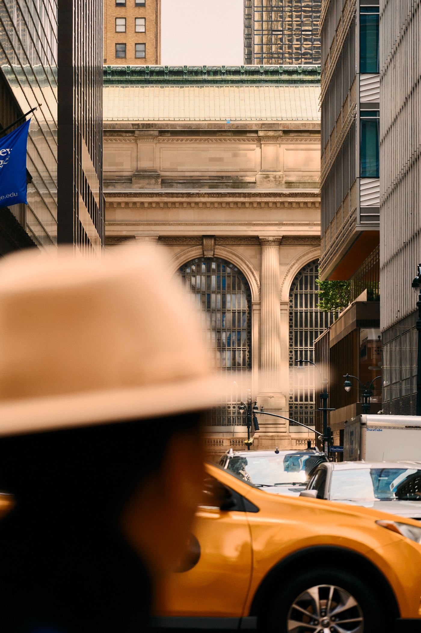 A bustling city street with tall buildings, a yellow taxi, and a person wearing a hat.