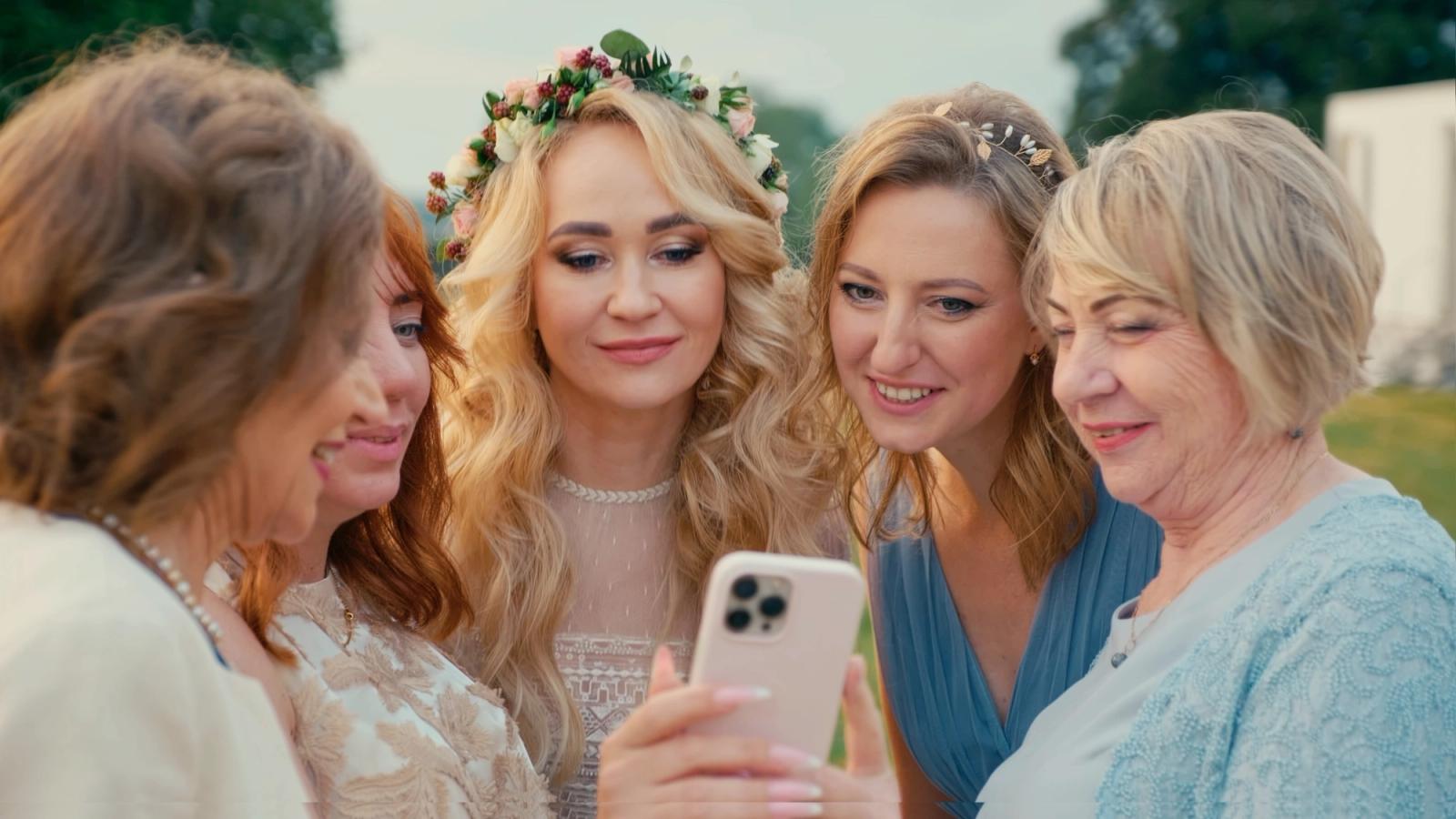 A group of women gather outdoors, with one woman in a white lace dress and a floral crown, taking a selfie with her phone.