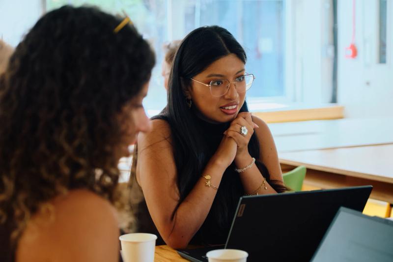 A woman with glasses and a black shirt is sitting at a table with a laptop.