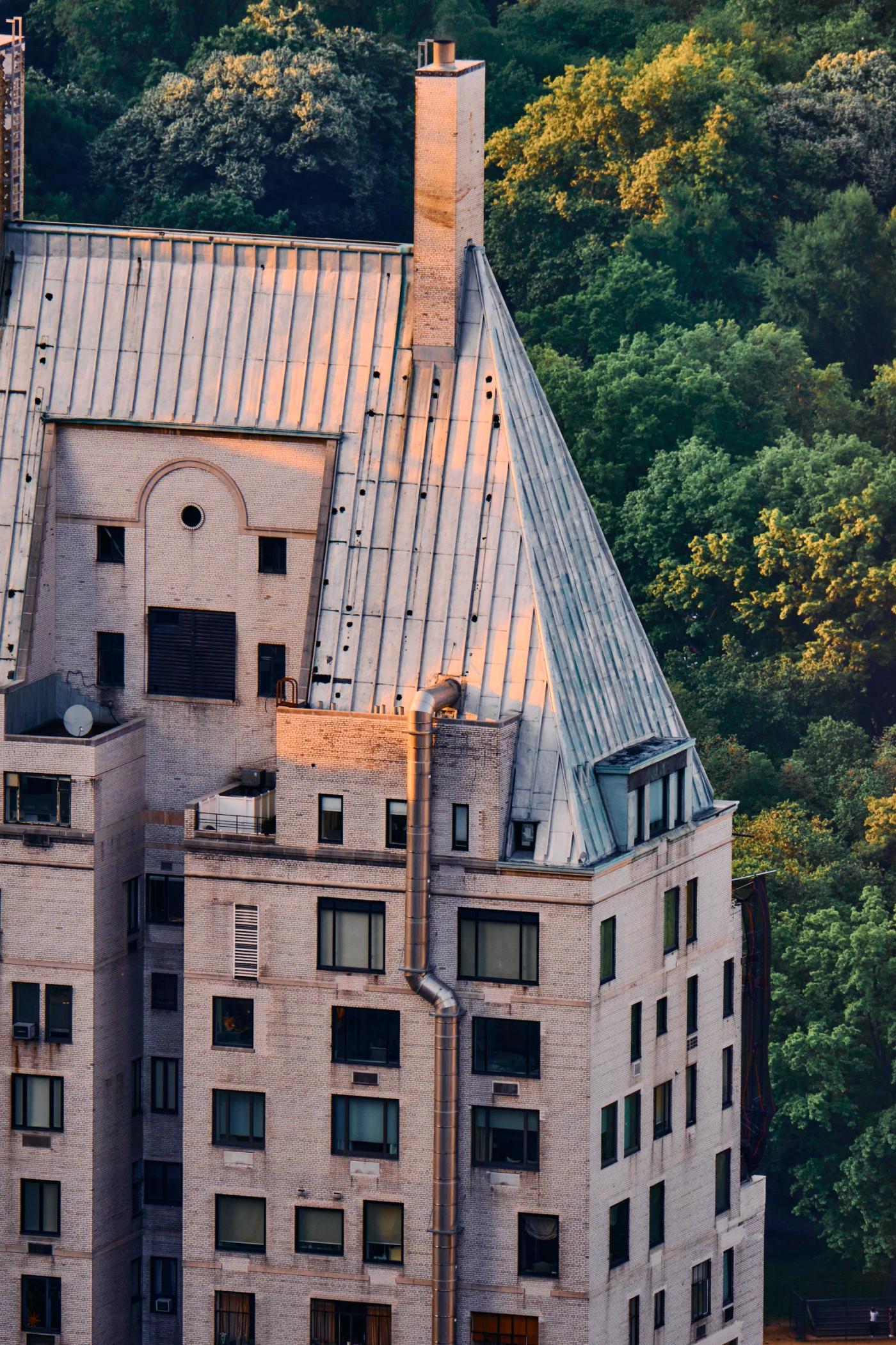 A large, multi-story building with a sloping roof and multiple chimneys is surrounded by lush green trees.