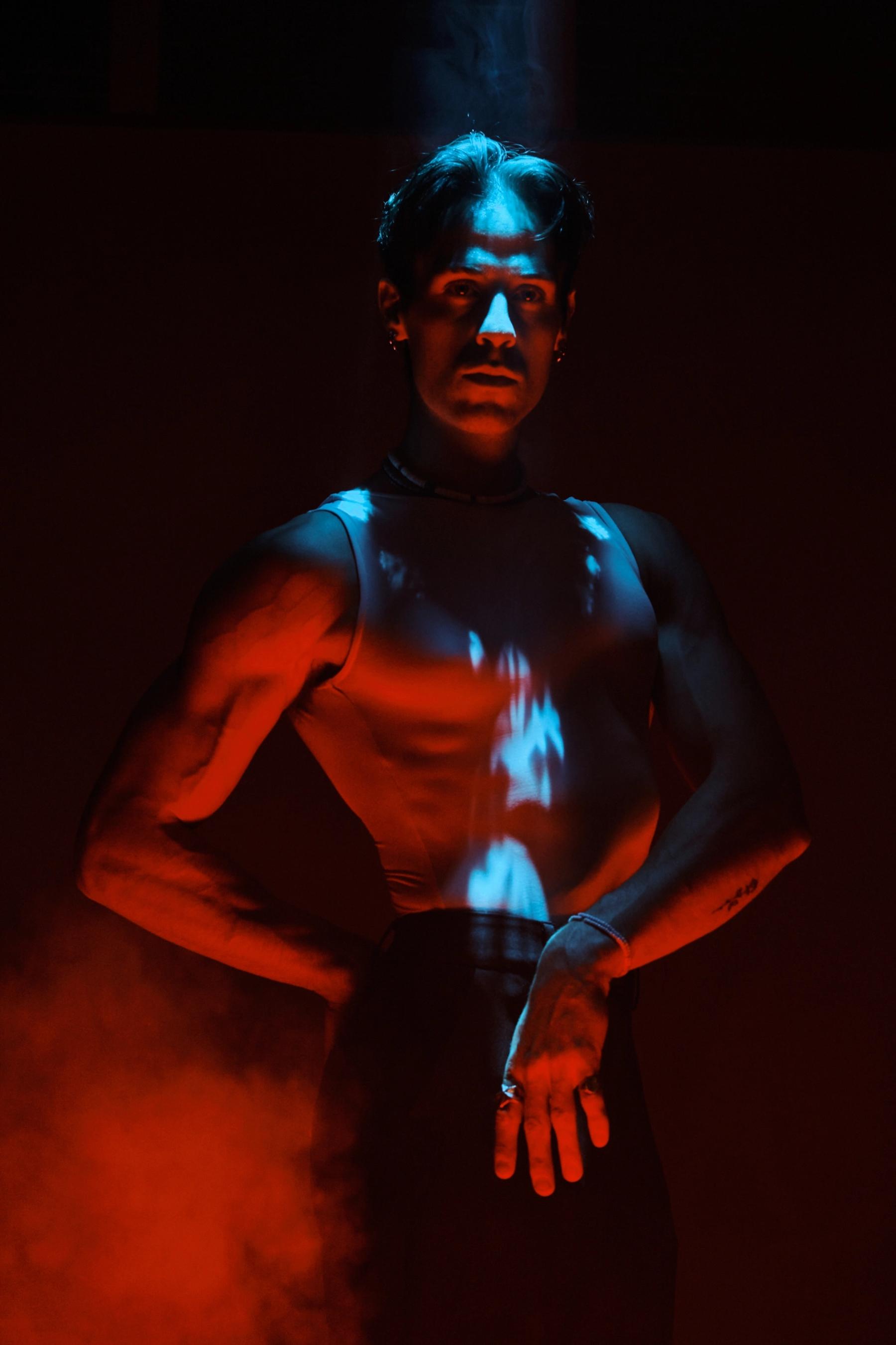 A man in a white tank top stands in a dimly lit room, with a red light illuminating his torso and hands. His hands are crossed over his chest, and he 