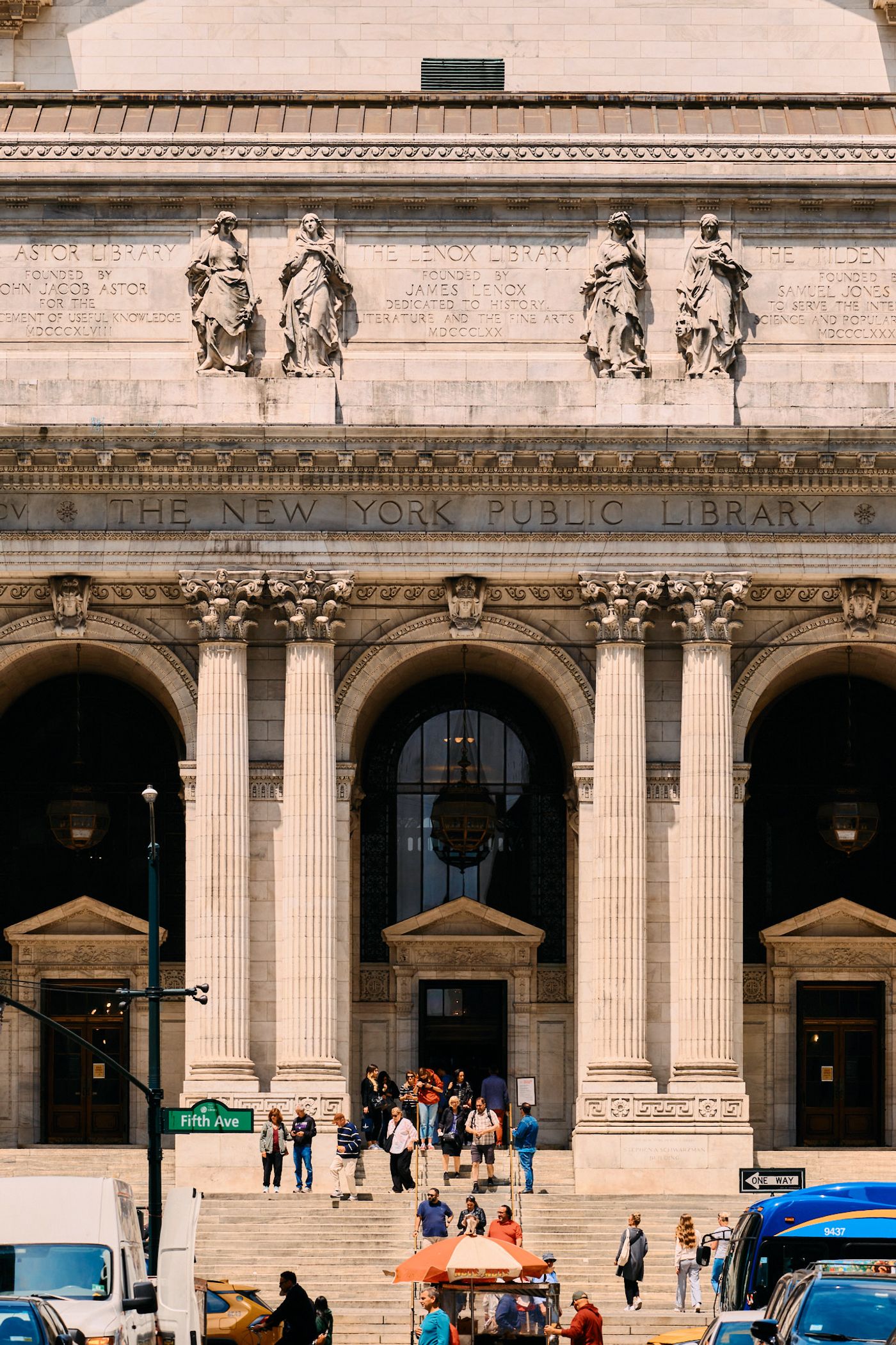 A bustling New York City street scene features a grand, ornate building with columns and statues, surrounded by pedestrians and vehicles.