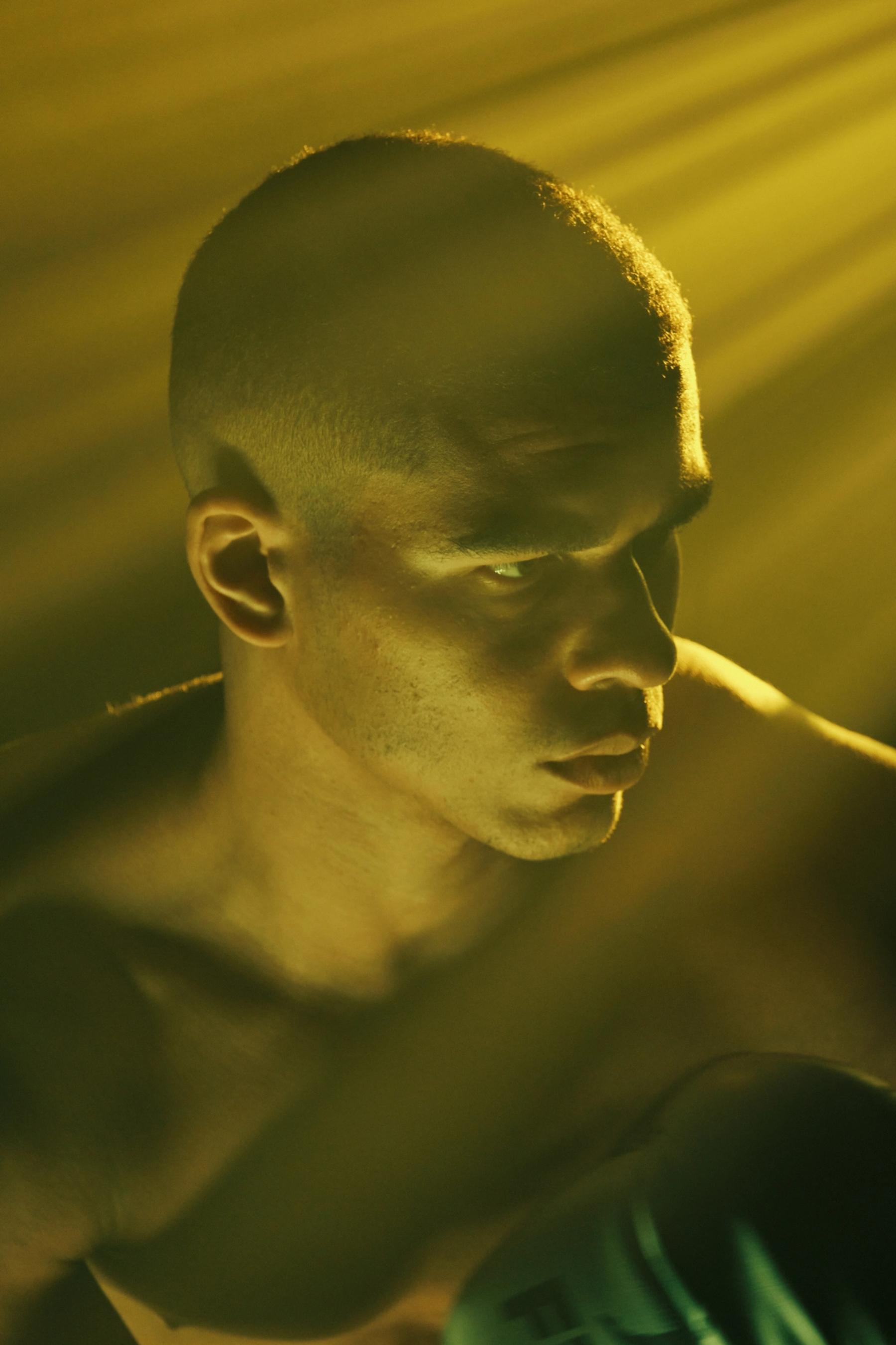 A muscular man in a black shirt stands in a dimly lit room, with a bright yellow light illuminating his face.