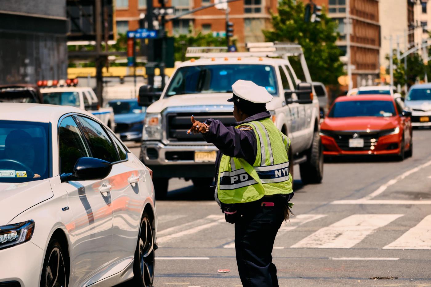 A police officer in a green vest and white hat is directing traffic on a busy city street.