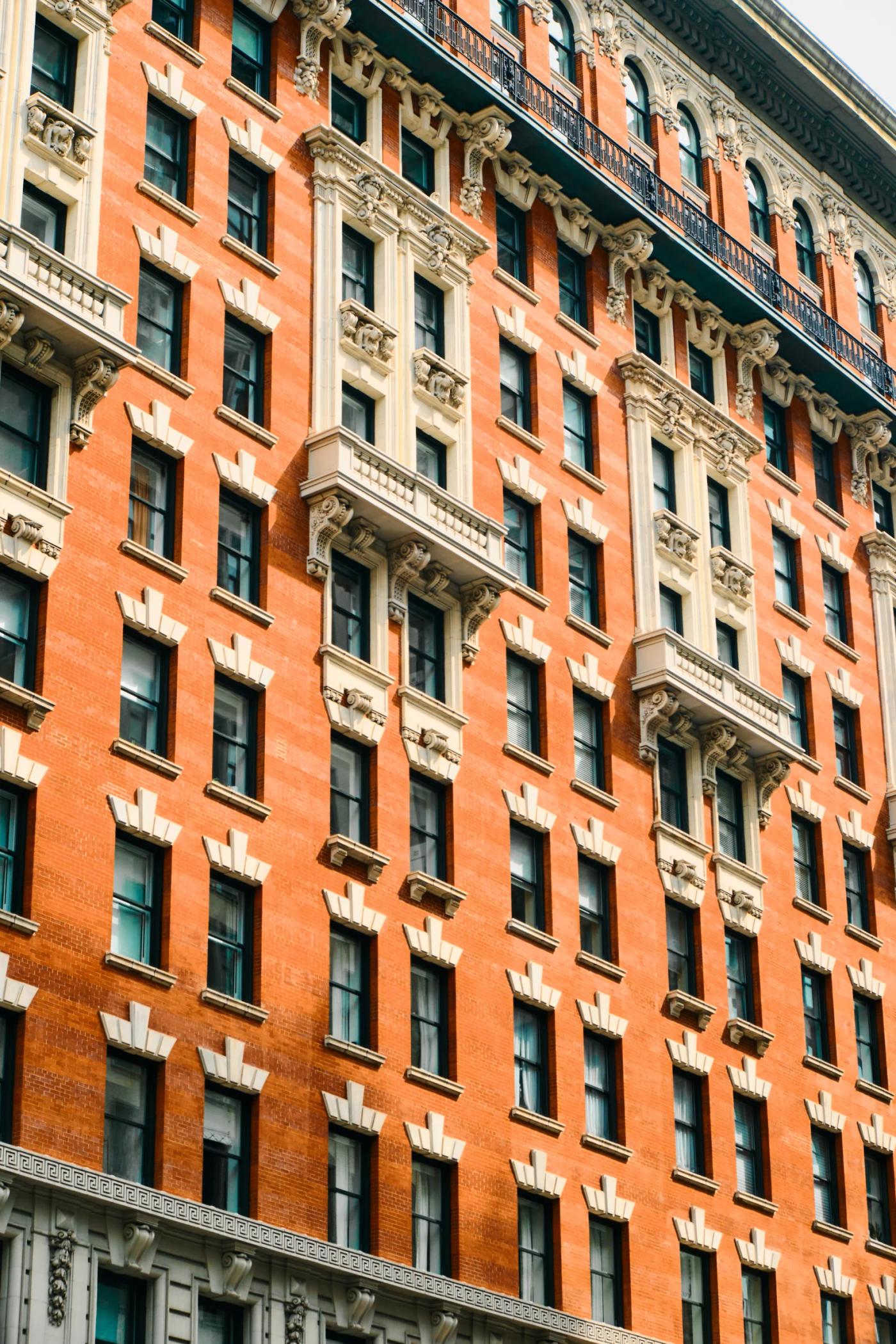 A red brick building with white trim and balconies stands tall in a bustling cityscape, with a clock tower nearby.