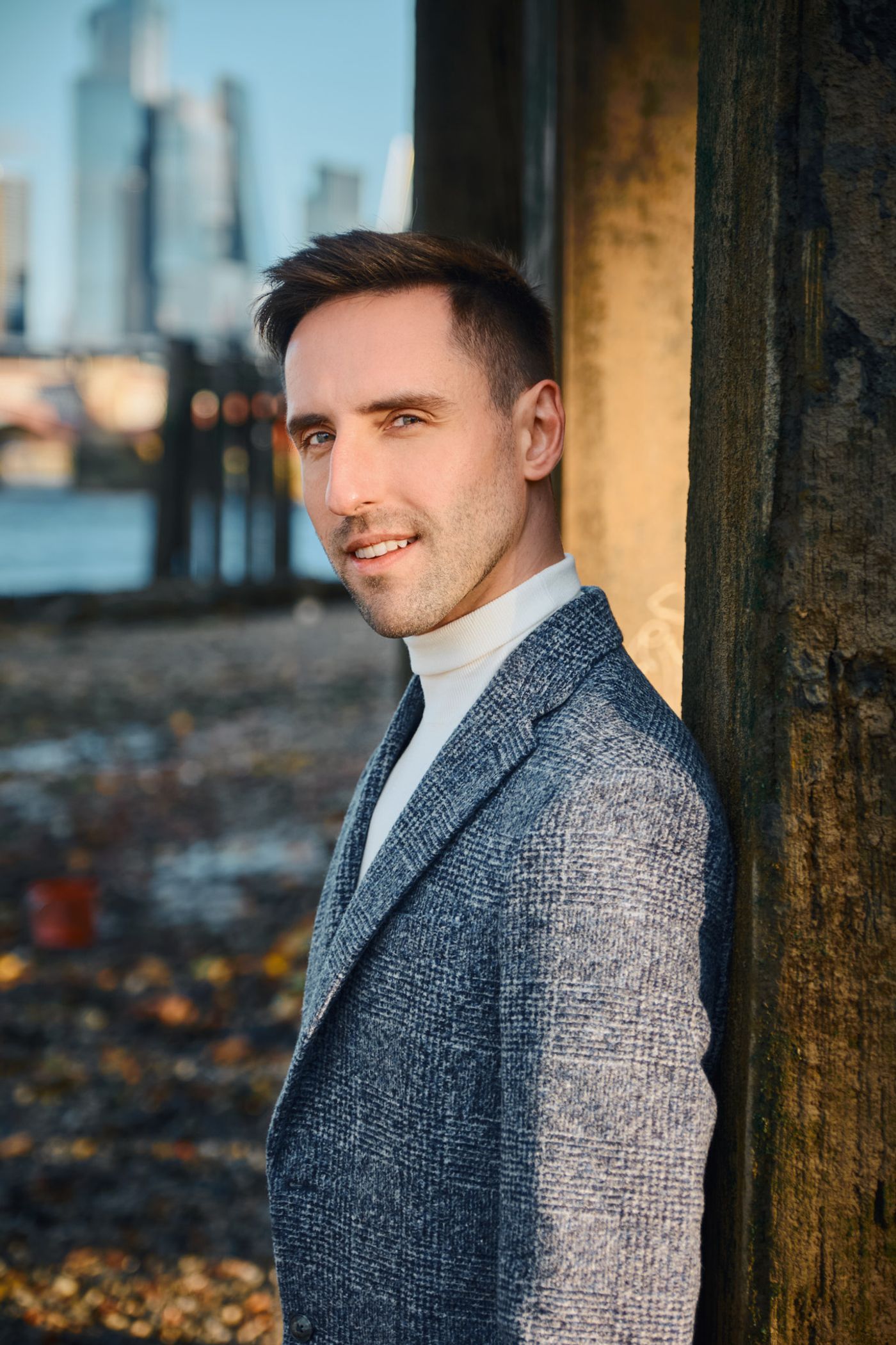 A young man in a gray blazer and white shirt stands confidently by a wooden pillar, gazing directly at the camera. The city skyline is visible in the 
