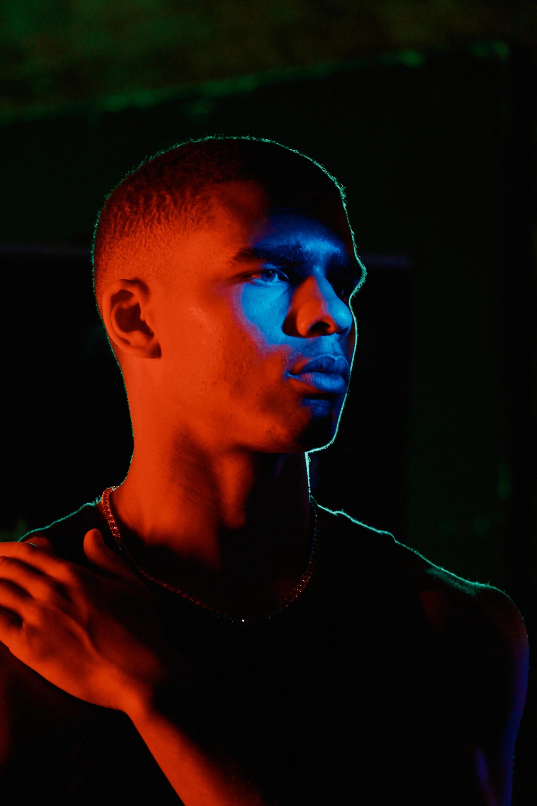 A young man in a black tank top, with a necklace, stands in a dimly lit room, gazing off to the side.