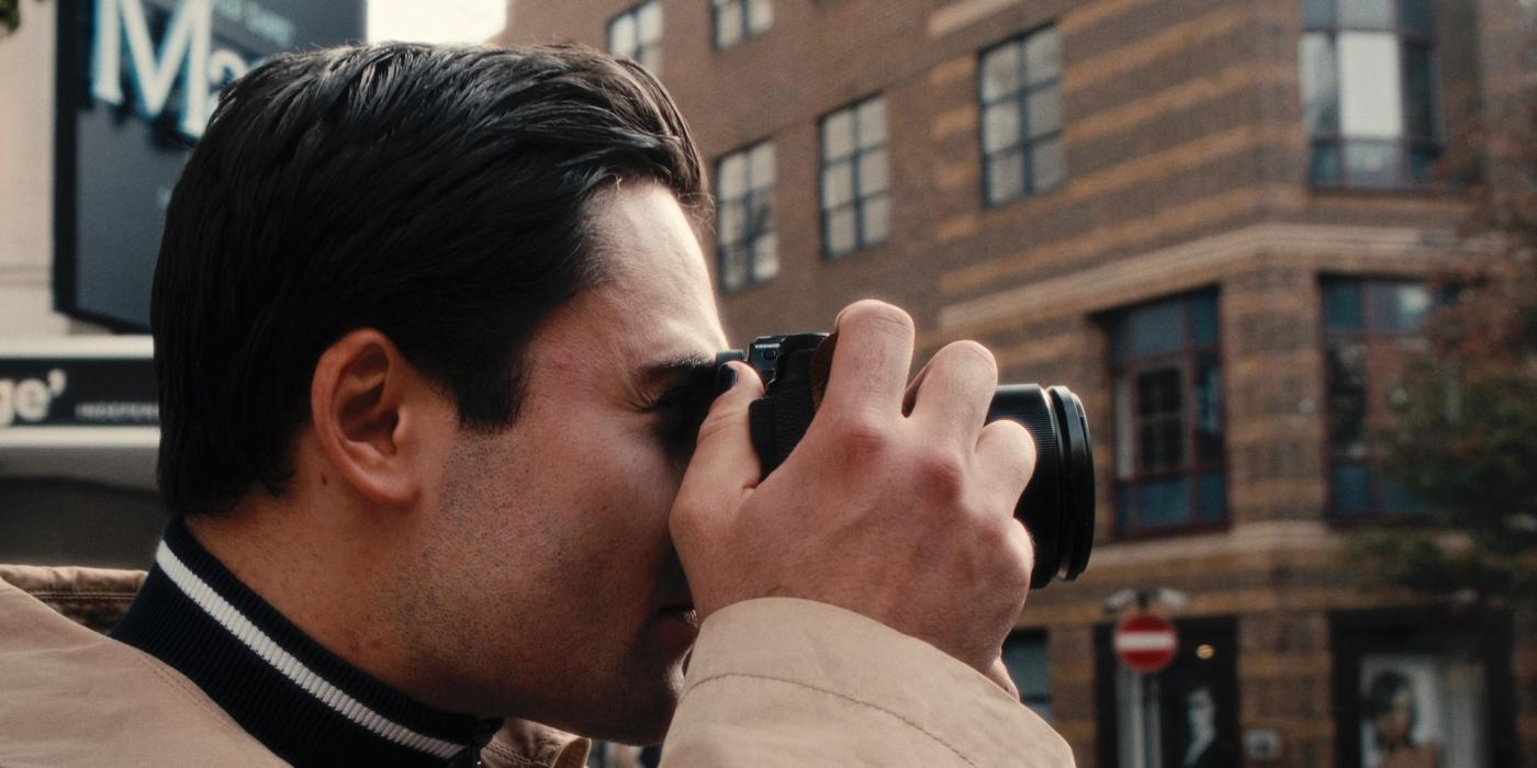A man in a beige jacket and black scarf is taking a photo with a black camera, focusing on the subject. He stands on a street with a brick building an