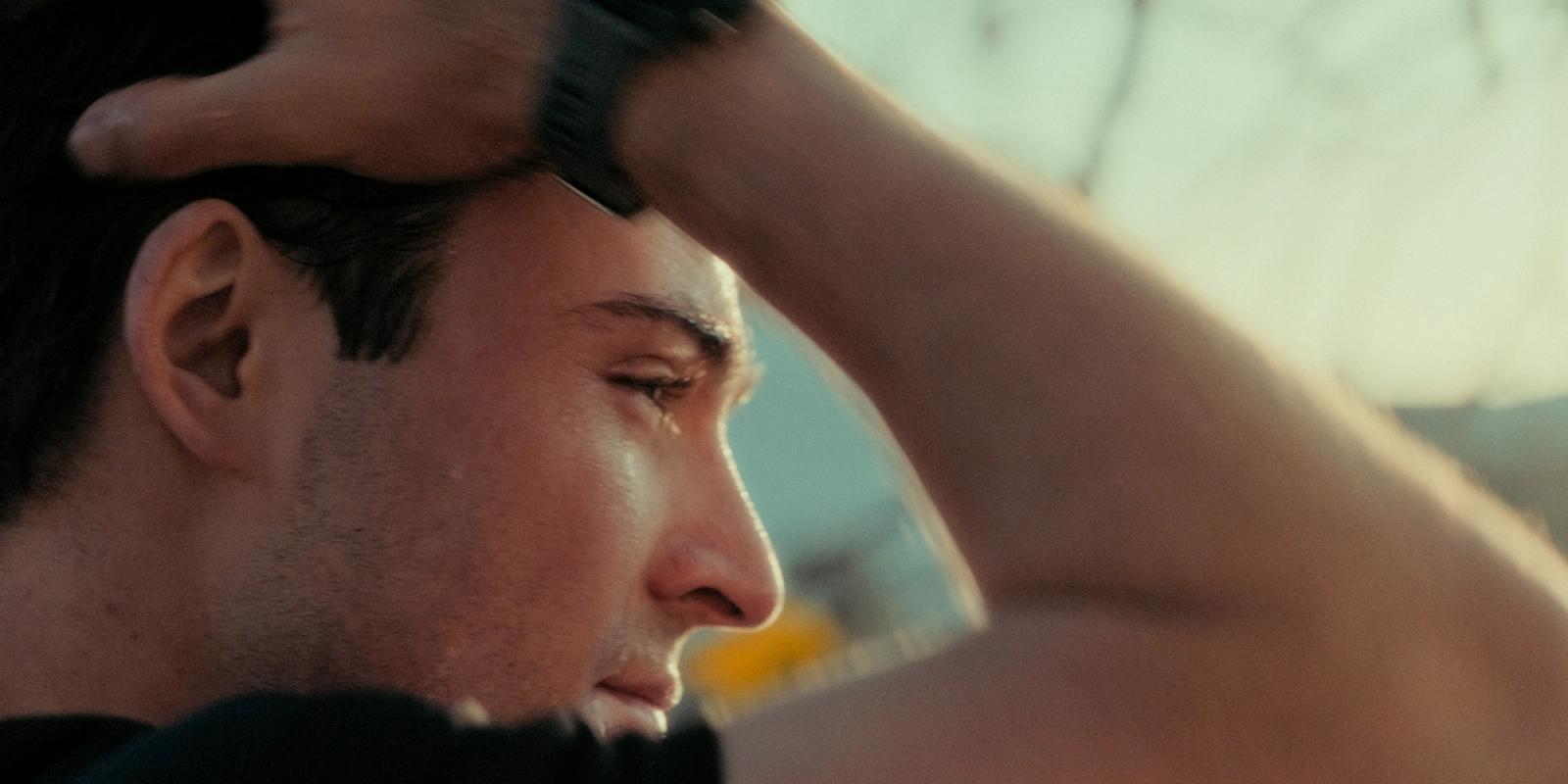 A close-up portrait of a man's face, with his head tilted to the side and hand on his head, against a blurred background of trees and a building.