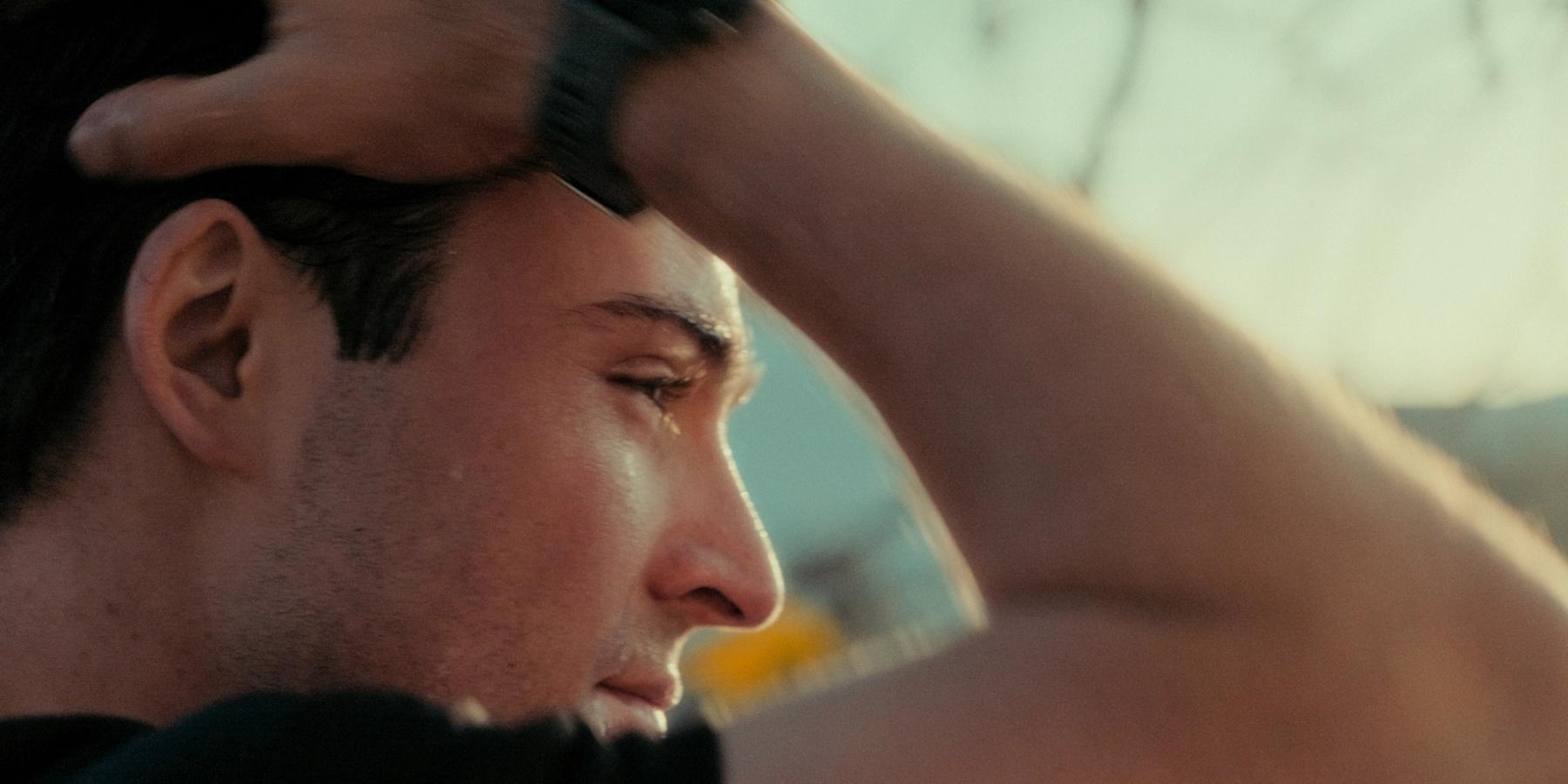 A close-up portrait of a man's face, with his head tilted to the side and hand on his head, against a blurred background of trees and a building.