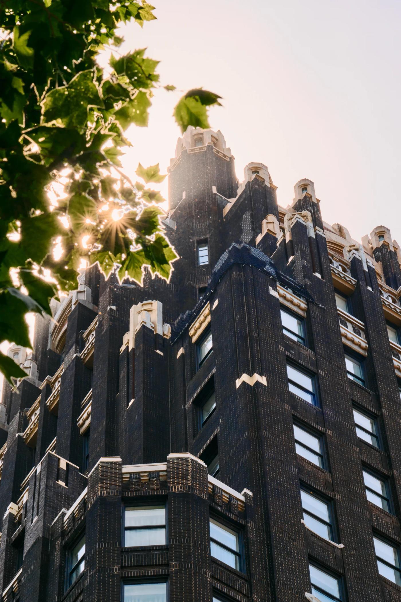 A tall, dark brick building with multiple levels and intricate architectural details stands in a lush green environment, with a tree on the left side 