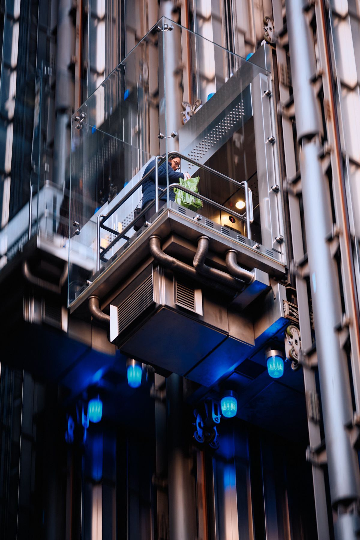 Contemporary glass and steel elevator shaft in narrow urban alleyway with blue LED accent lighting and silhouetted figure at dusk