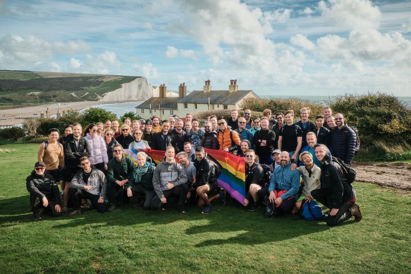 A group of people gather on a grassy hill, posing for a photo with a rainbow flag. The white cliff houses and ocean are visible in the background.
