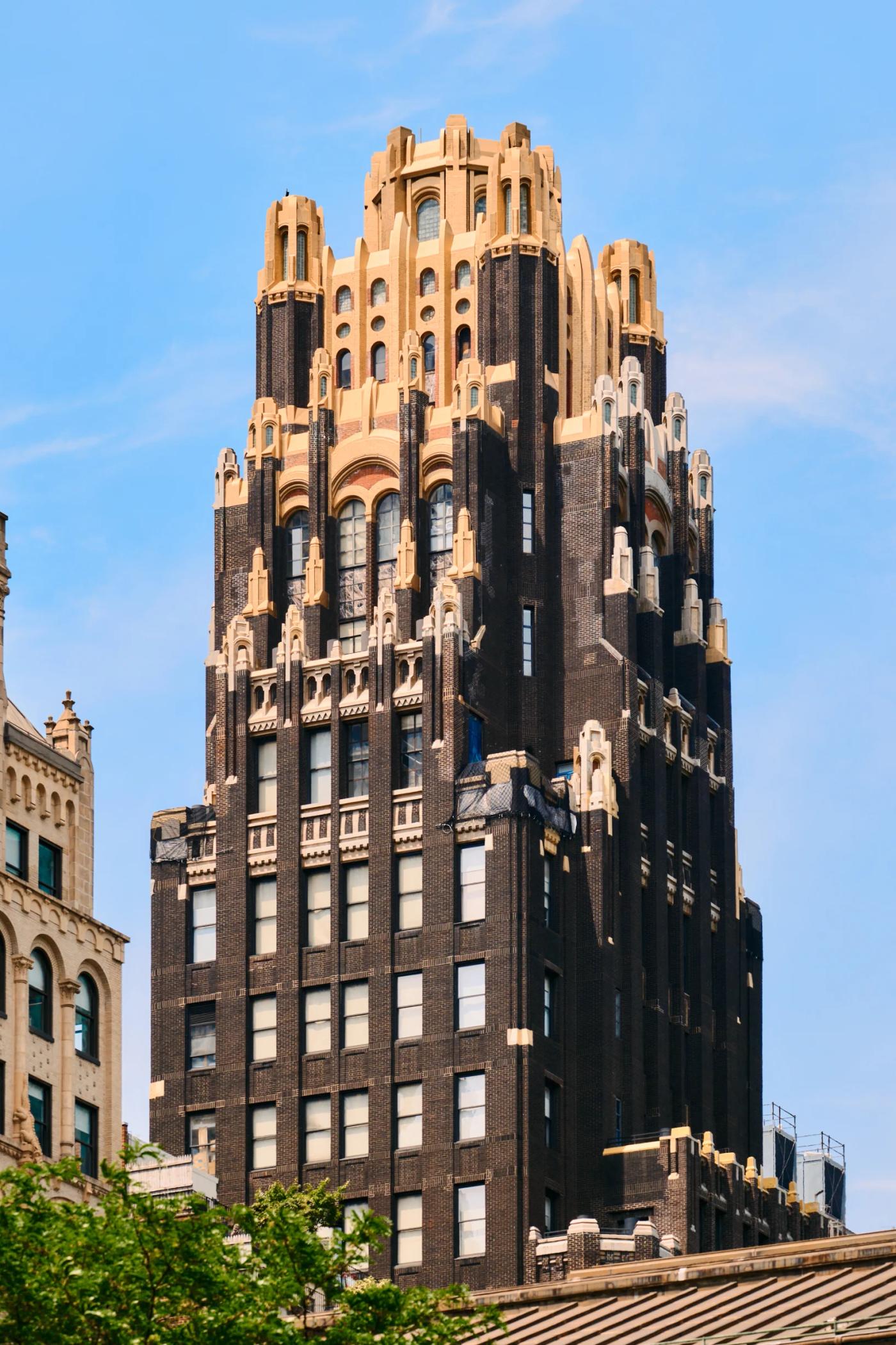A tall, dark brown building with ornate spires and a golden-topped spire stands in a bustling cityscape, surrounded by other buildings and trees.
