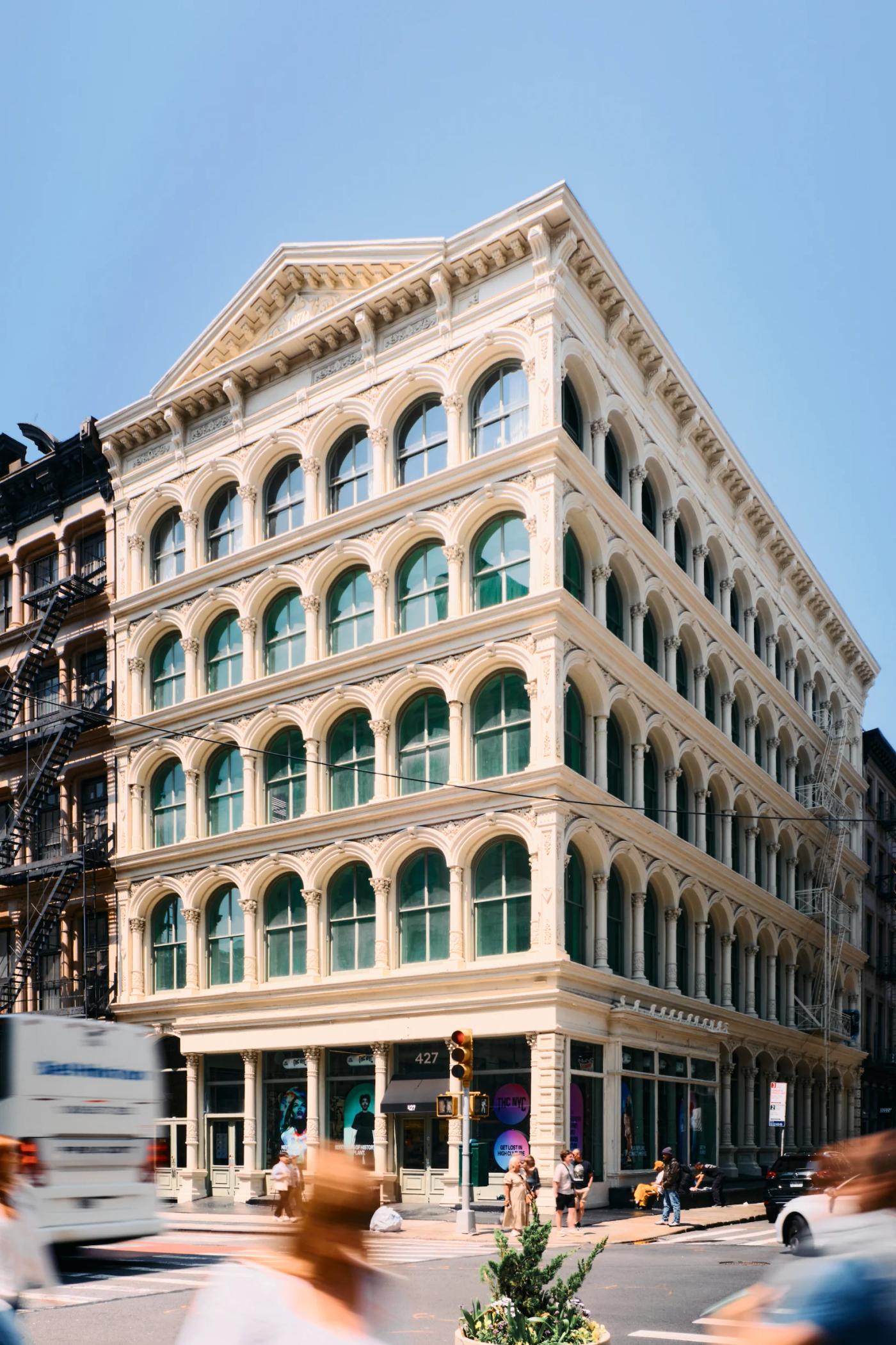 A white, ornate building with green shutters stands on a bustling city street, surrounded by other buildings and cars.