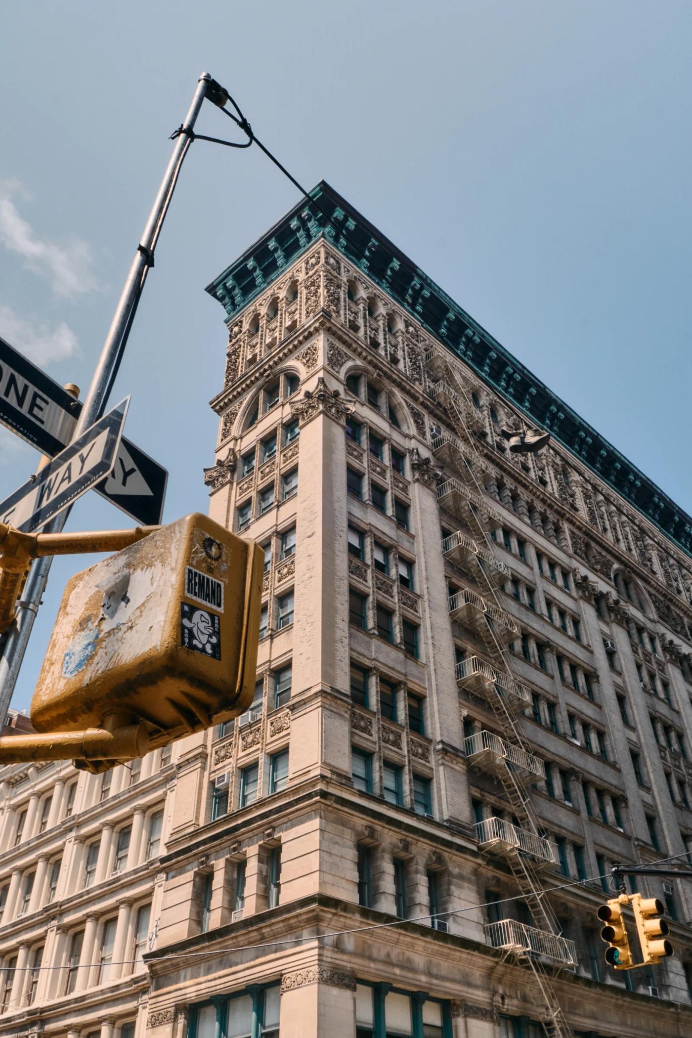 A tall, beige building with intricate carvings and a green fire escape stands in a bustling city corner. A yellow traffic light and a black street sig