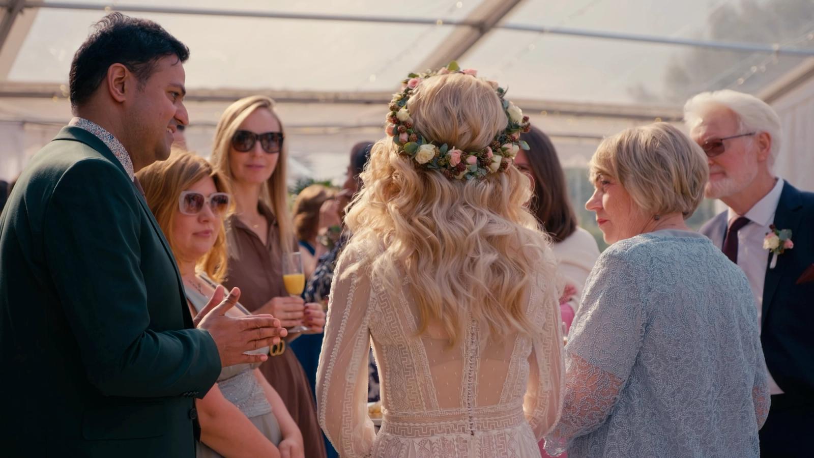 A wedding reception is captured in a tent, with a bride in a white dress and a man in a green suit holding a glass of orange juice. The group of peopl