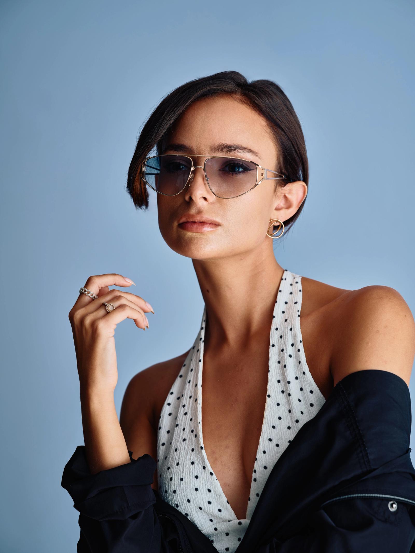 A stylish young woman with short, dark hair and a black jacket with polka dots is posing for a portrait against a blue background. She wears sunglasse