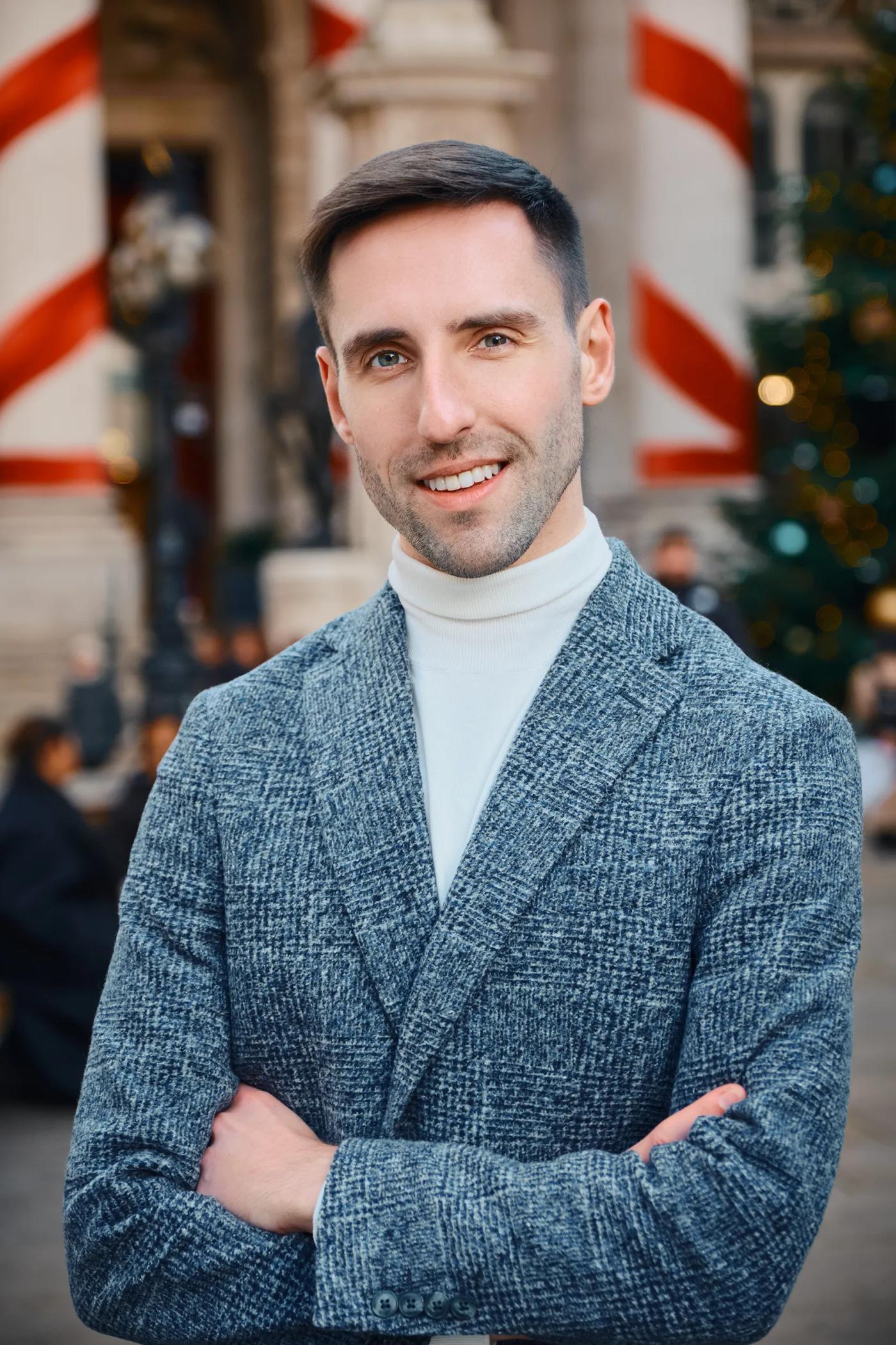 A man in a gray blazer and white turtleneck stands confidently in front of a Christmas tree, smiling at the camera.
