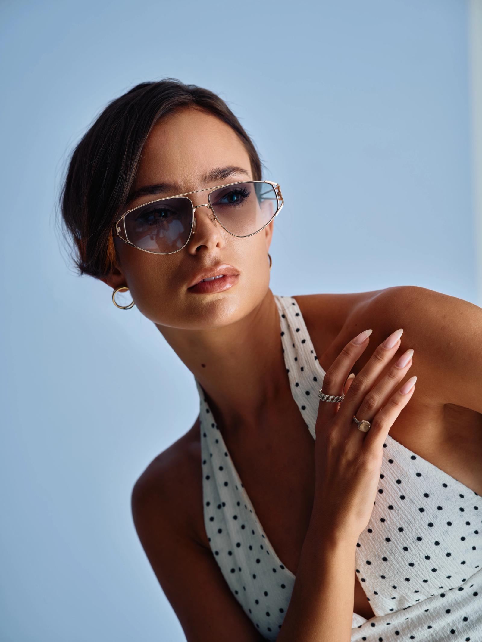 A young woman in a polka dot dress and silver sunglasses poses elegantly against a light blue background.