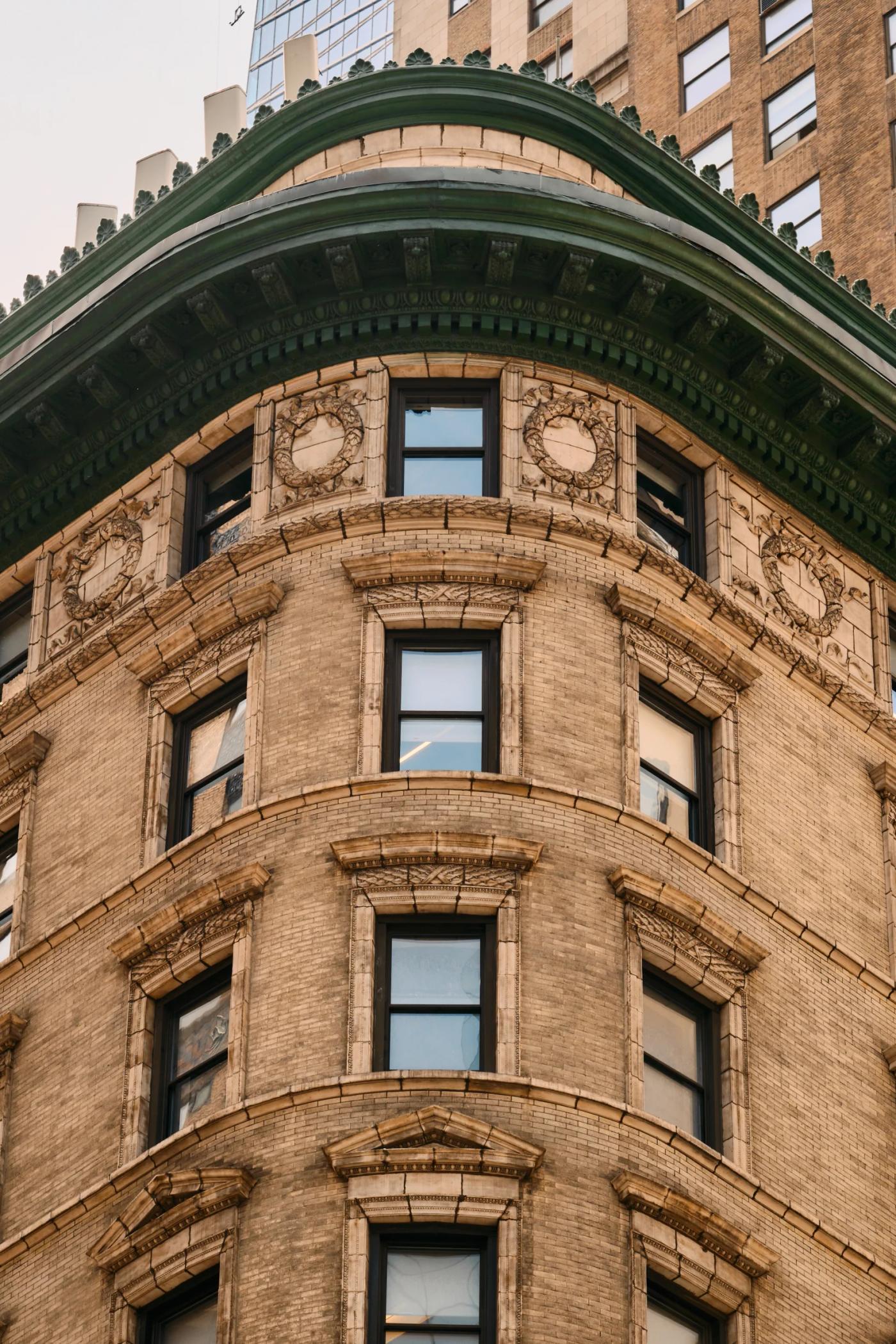 A tall, brown brick building with a curved facade and green trim stands in a bustling city, surrounded by other buildings and a clear sky.