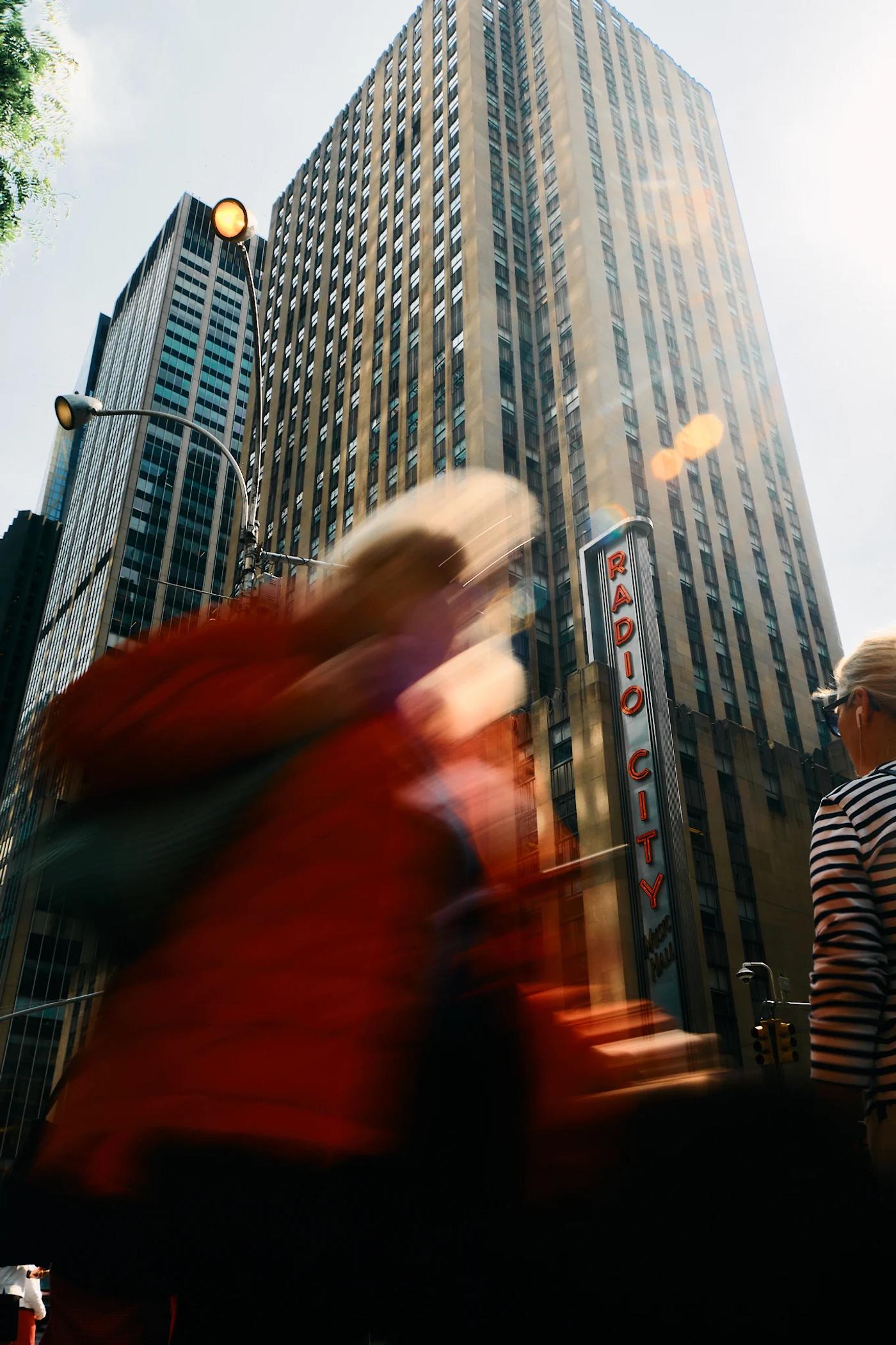 A bustling city street with tall buildings, a red jacketed person, and a blurred person walking.