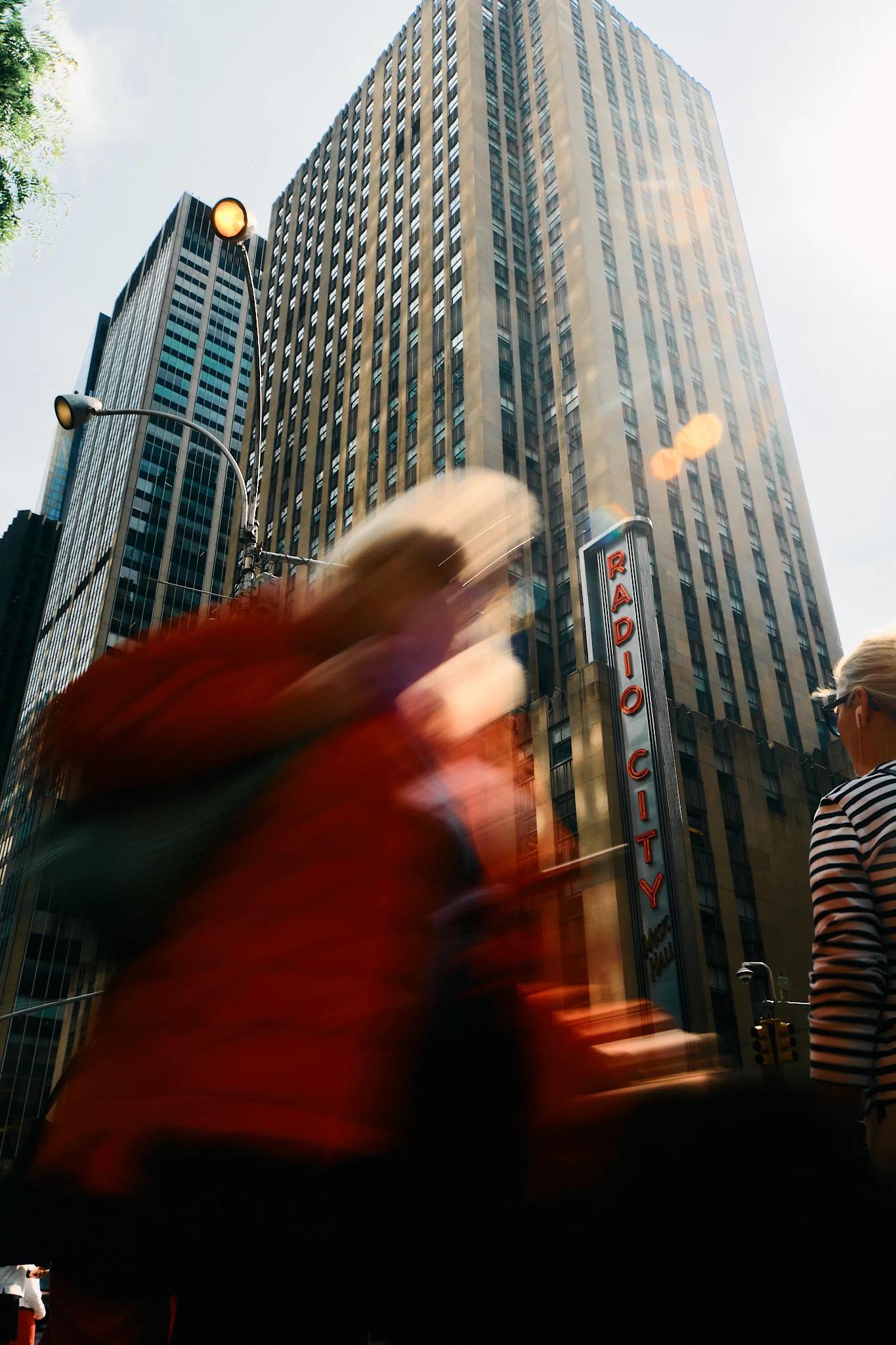 A bustling city street with tall buildings, a red jacketed person, and a blurred person walking.