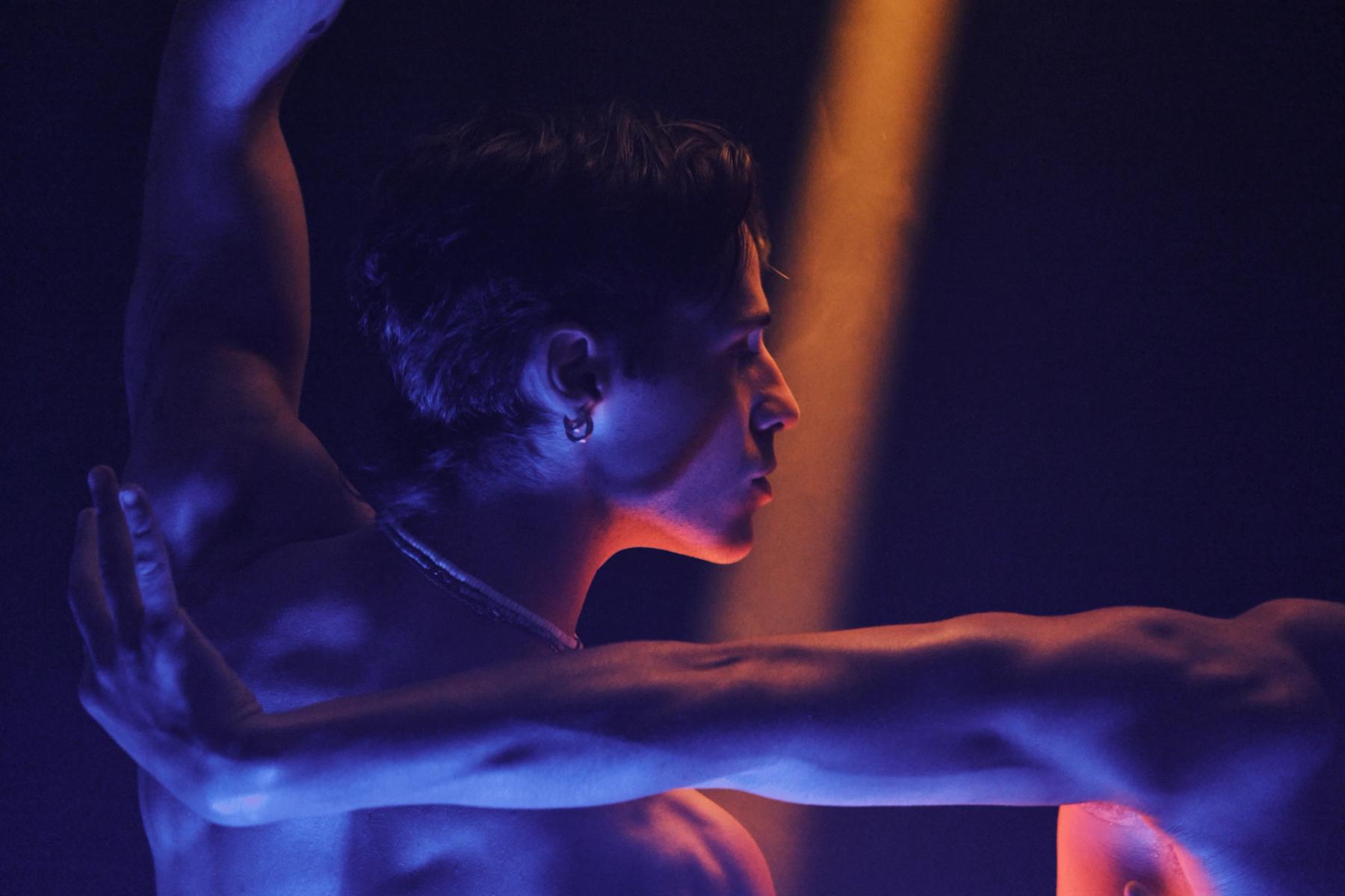 A shirtless dancer in a blue and purple light, with a necklace, is captured in a dramatic pose on a stage, illuminated by a spotlight.