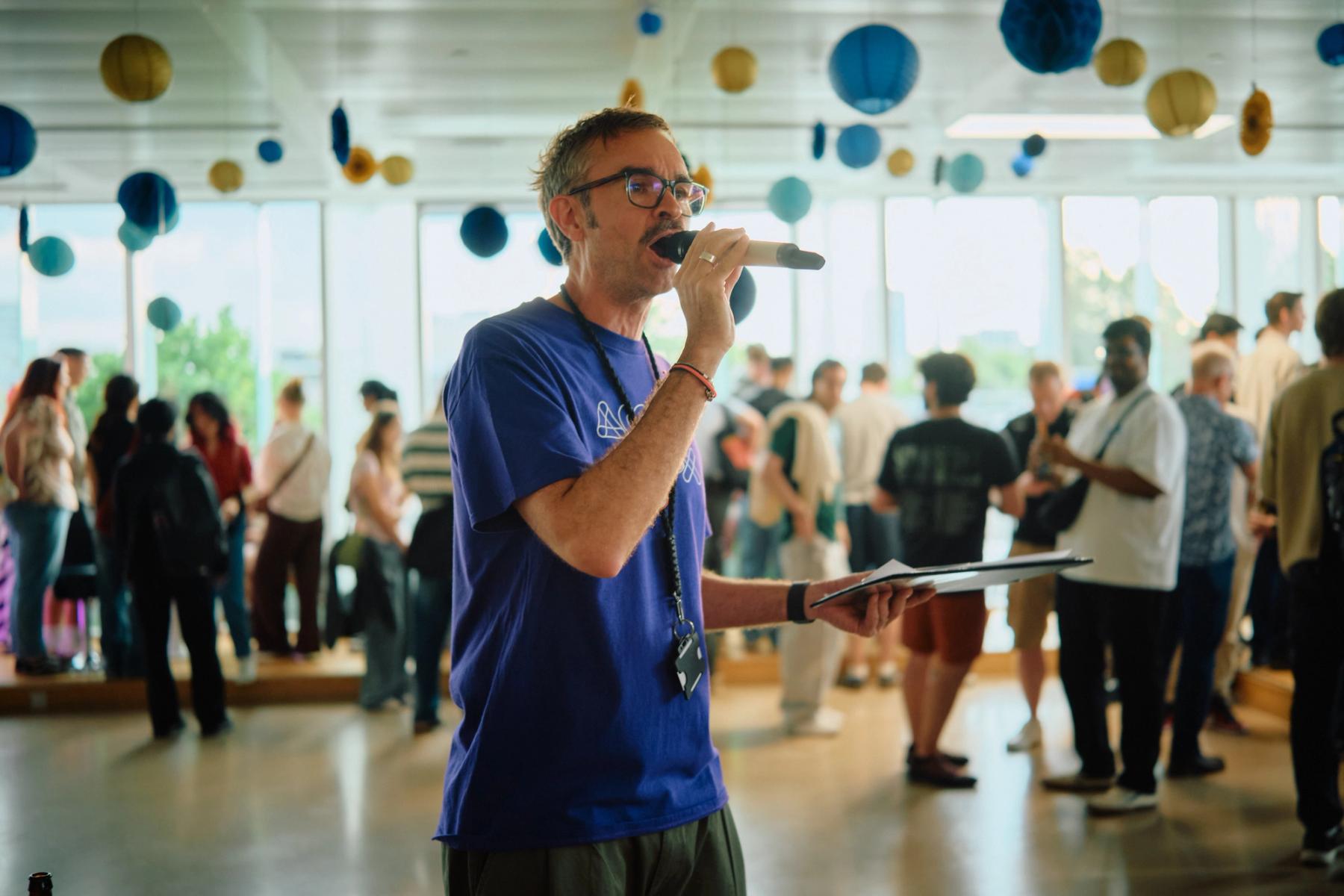 Access VFX community event 2026, host in a purple shirt speaking into a microphone with blue and gold lanterns overhead