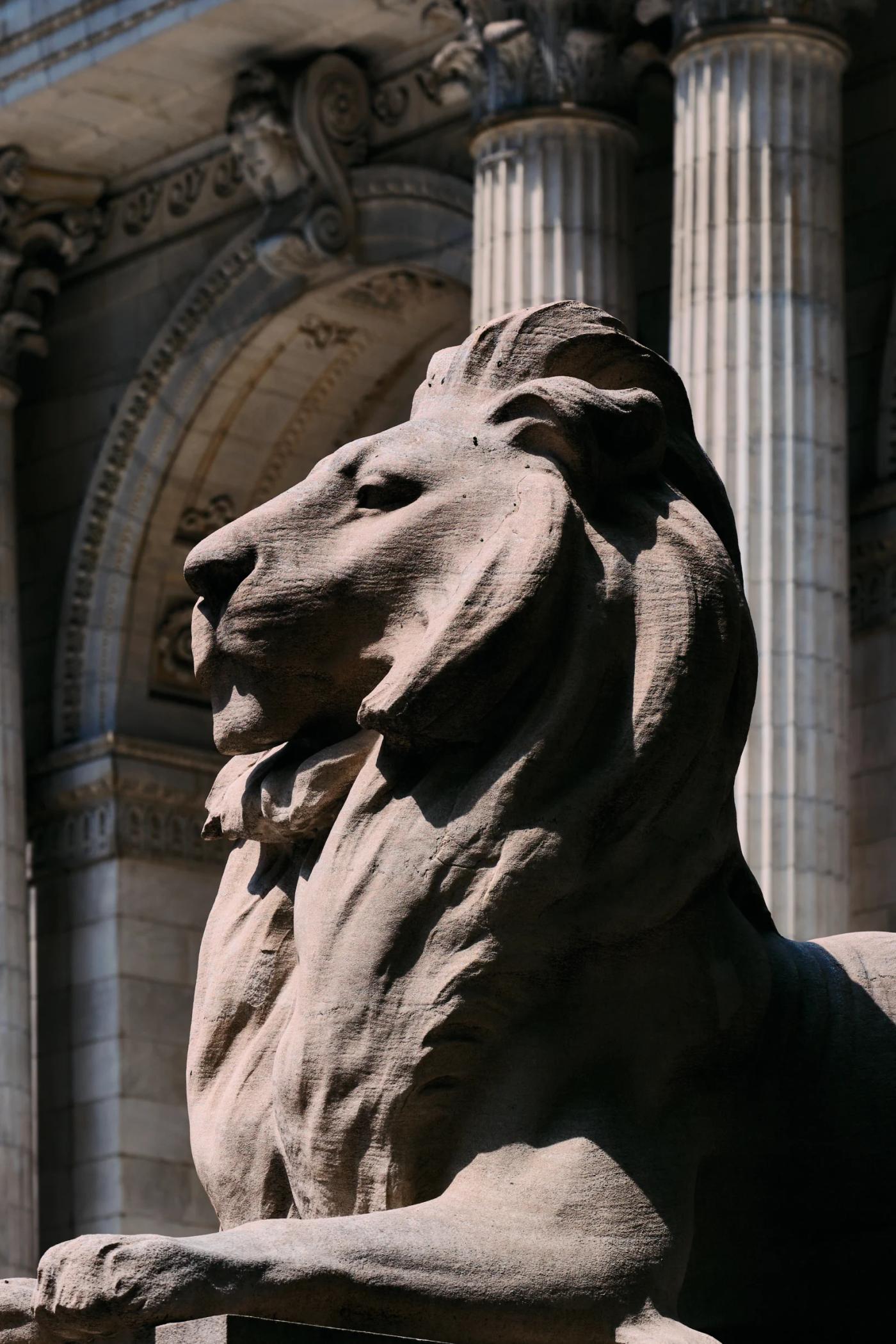 A majestic lion statue stands tall in front of a grand building with ornate columns, gazing to the left.