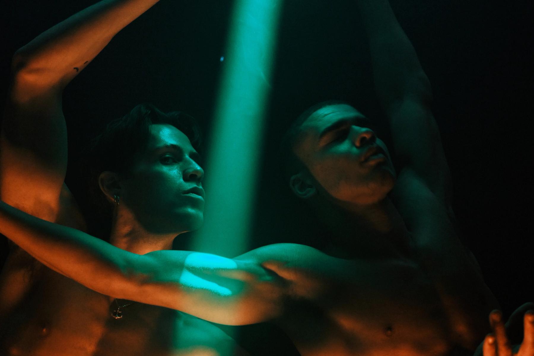 Two shirtless men in a dark room, one reaching upwards and the other reaching downwards, illuminated by a green light.
