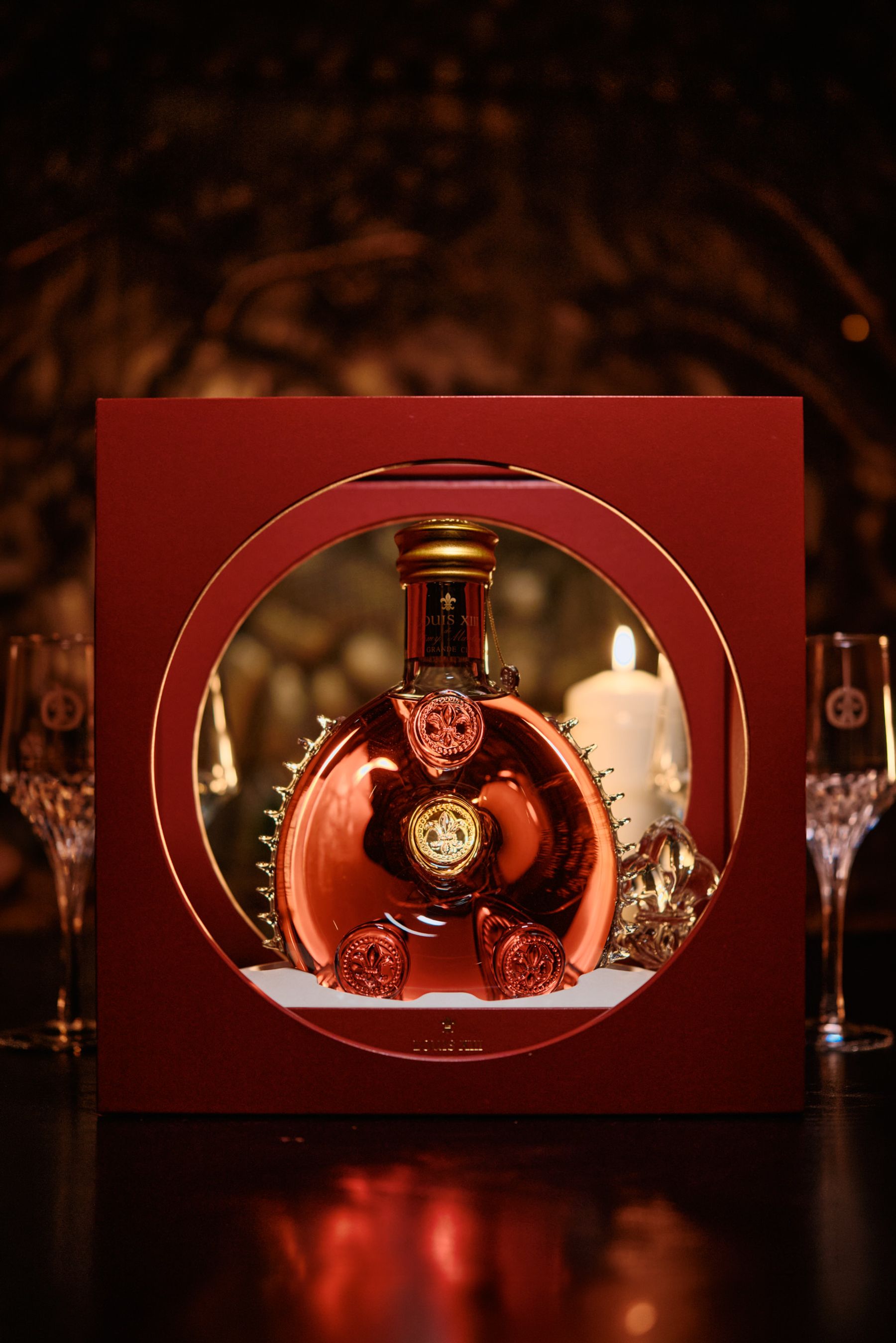 Louis XIII Cognac A red box with a gold cap and rose decoration is placed on a table, with a candle in the background. The box contains a red bottle with a gold cap and