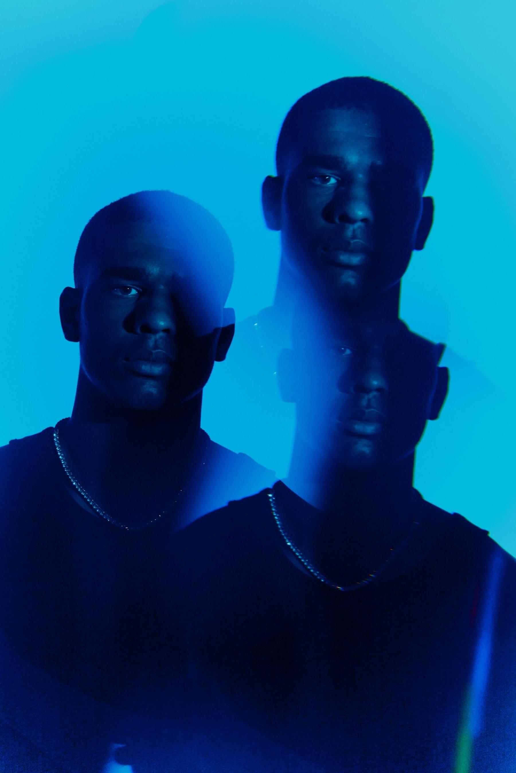 Two men in black shirts, one on the left and one on the right, with blue backgrounds, appear to be in a portrait.