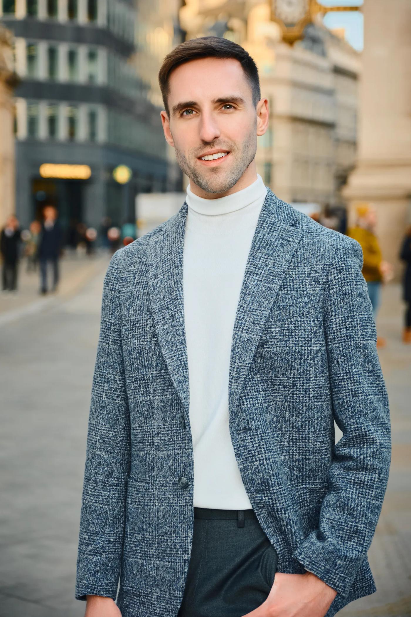 A man in a gray blazer, white turtleneck, and white sweater stands on a city street, posing for a photo.