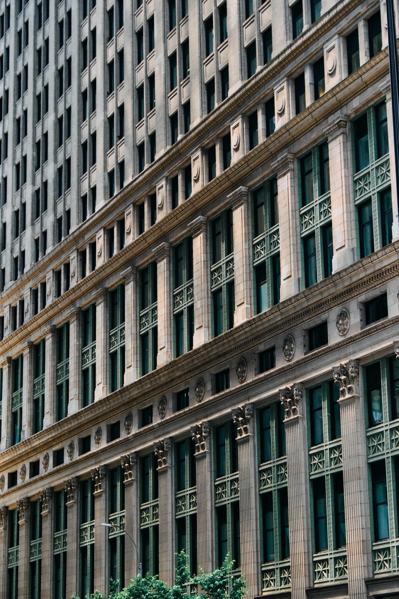 A tall, modern building with many windows and green shutters stands in a bustling cityscape, with a pedestrian crossing in the foreground.