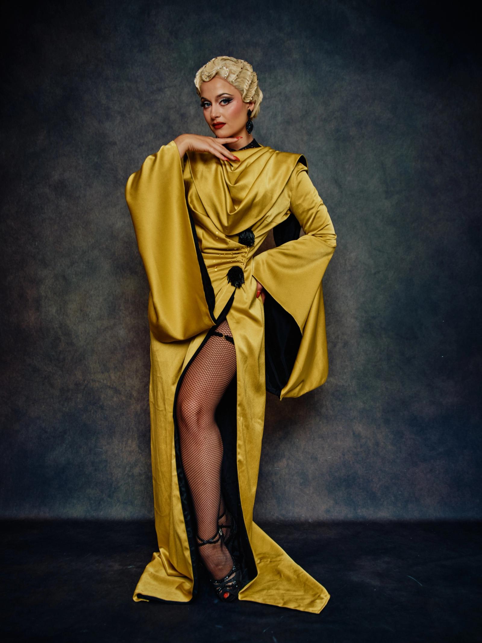 A woman in a golden dress with a black trim and a black belt stands confidently against a dark background. Her blonde hair is styled in a bob, and she