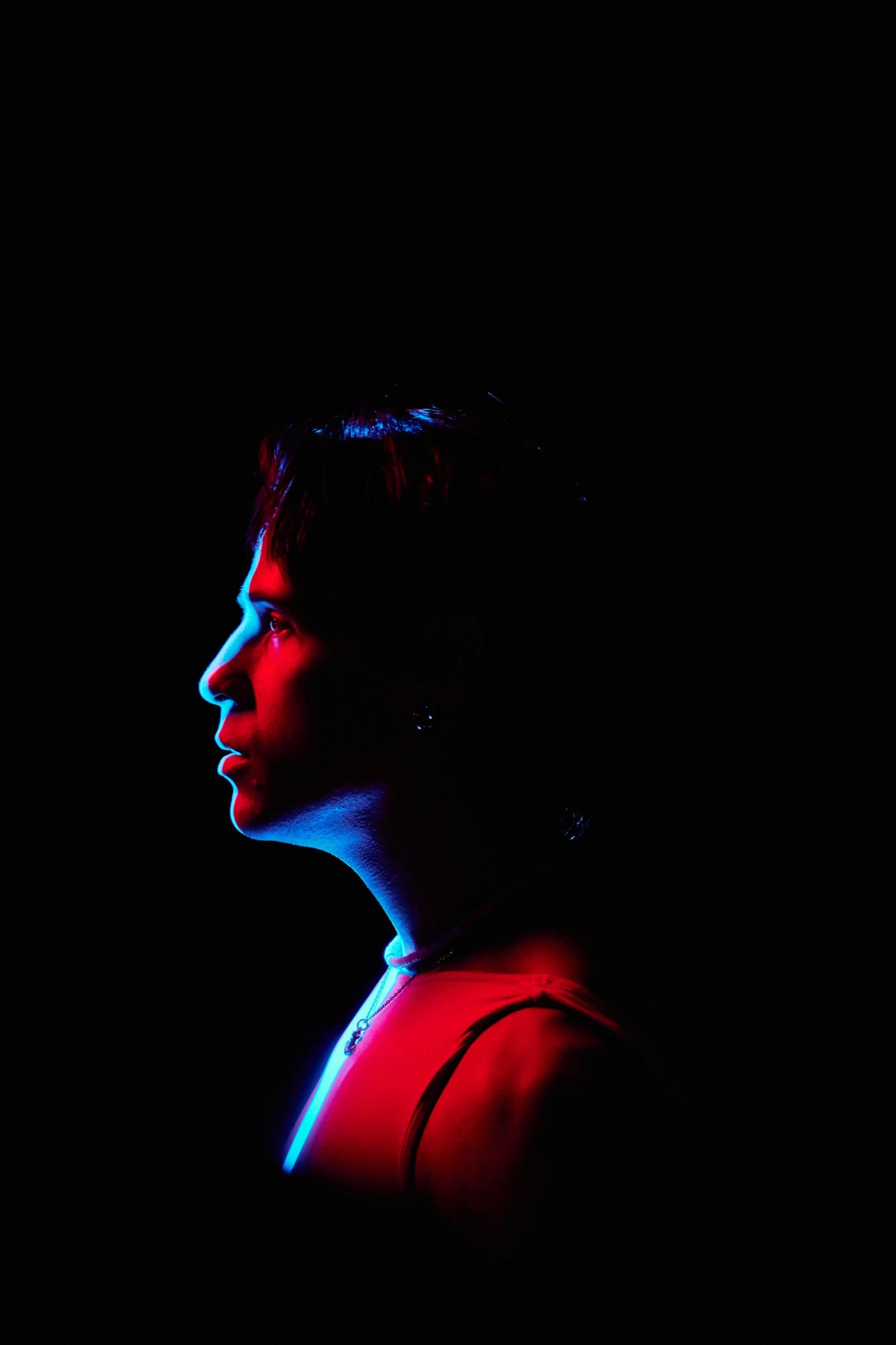 A woman in a red dress, with a red nose and red hair, is illuminated by blue light against a black background.