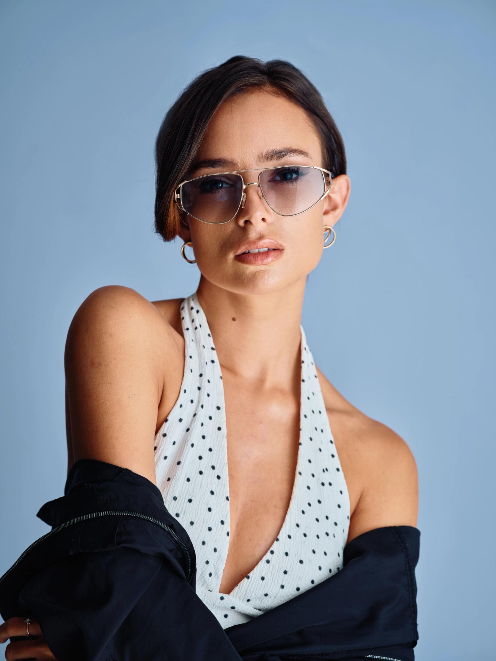 A young woman with short, dark hair and a stylish hairstyle wears sunglasses and a polka dot tank top, with a black jacket over her shoulder. She gaze