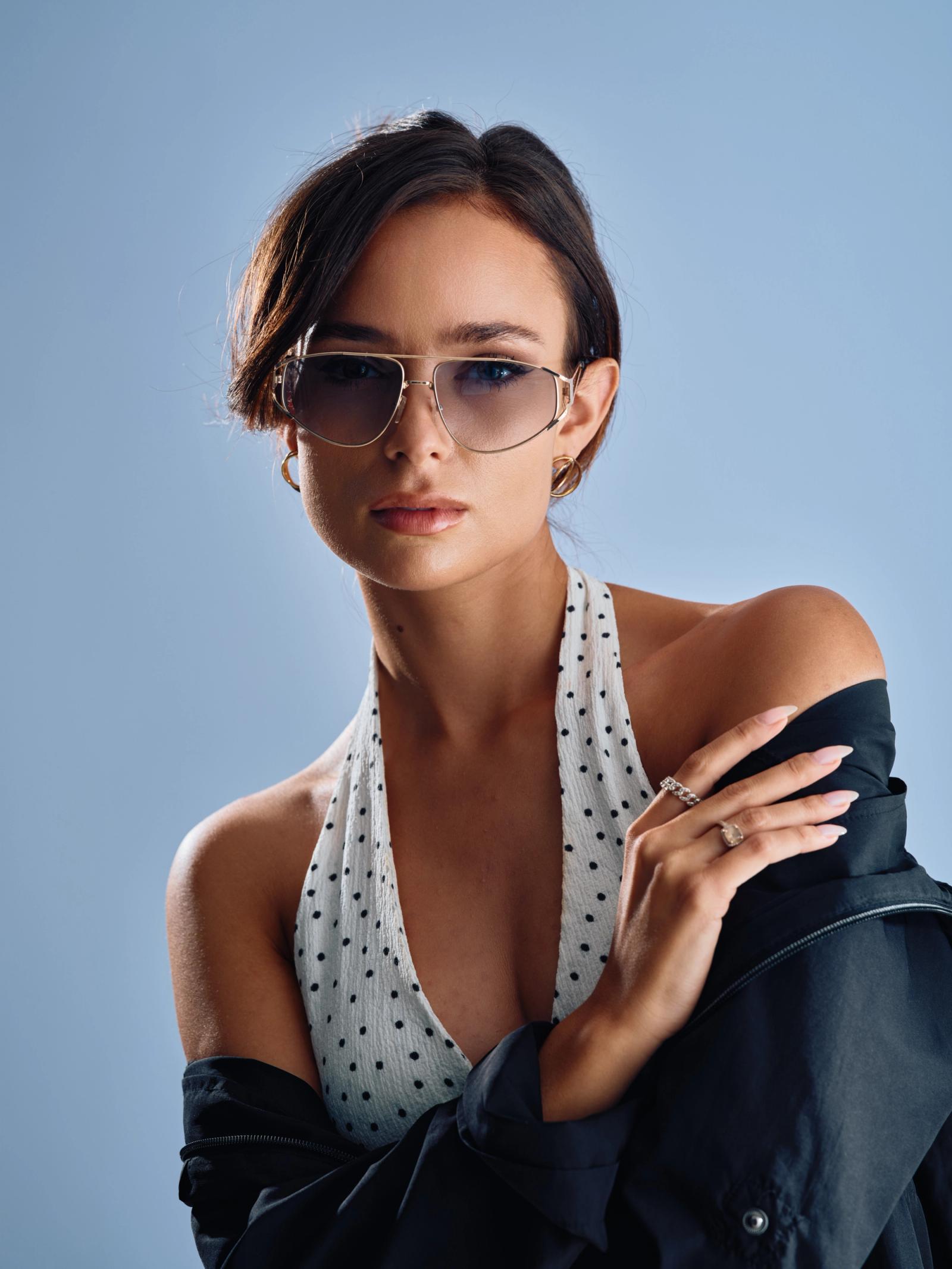 A stylish young woman in a white polka dot dress and black jacket, wearing sunglasses, poses confidently with her hands on her hips and a serious expr