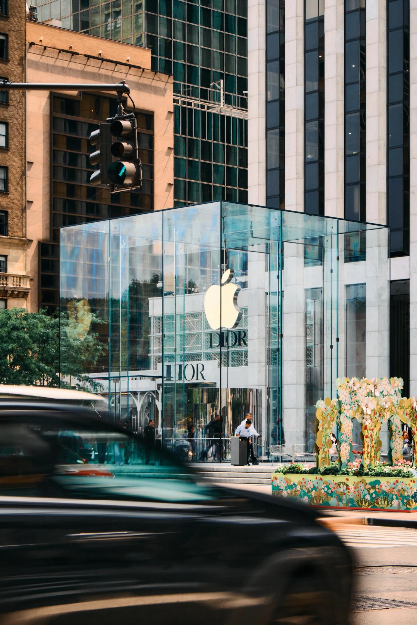 A bustling city street scene features a large Apple store with a glass facade, surrounded by tall buildings and a traffic light. Cars are parked on th