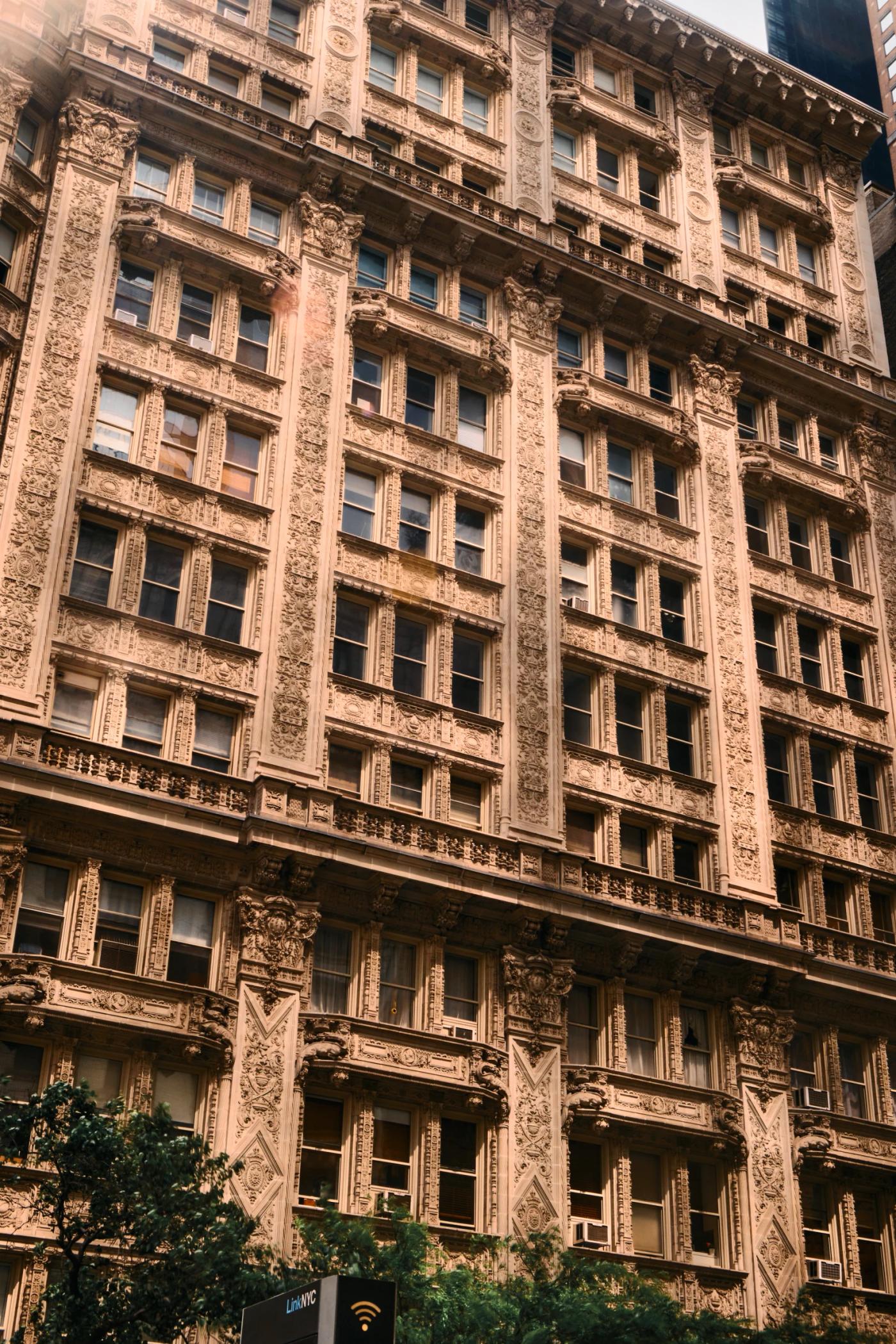 A tall, beige stone building with intricate carvings and columns stands in a bustling city, surrounded by trees and other buildings.