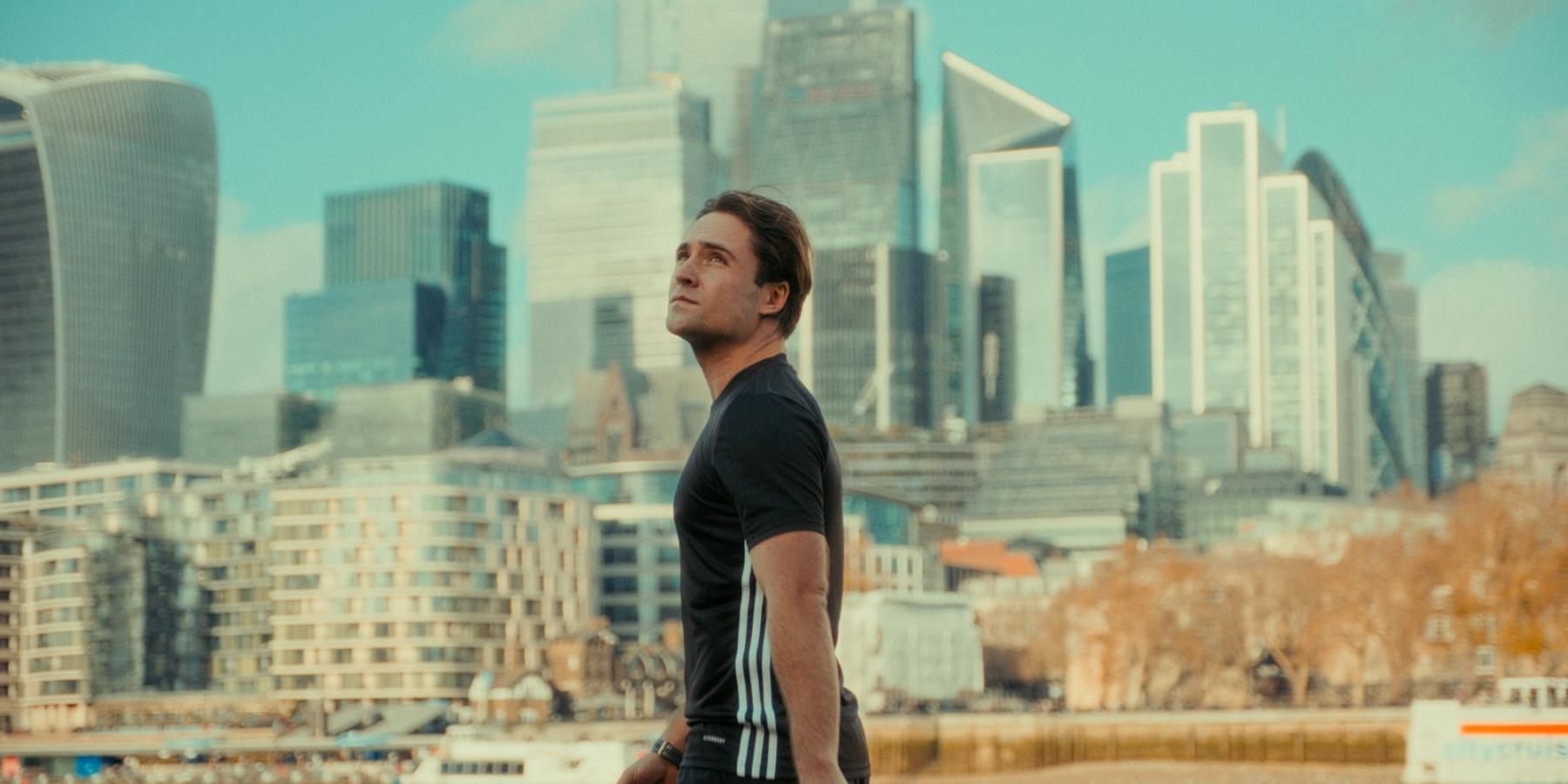 Max Stone, actor, stands on a riverbank, wearing a black t-shirt with white stripes and a black belt. He gazes upwards, gazing at the city skyline. Th
