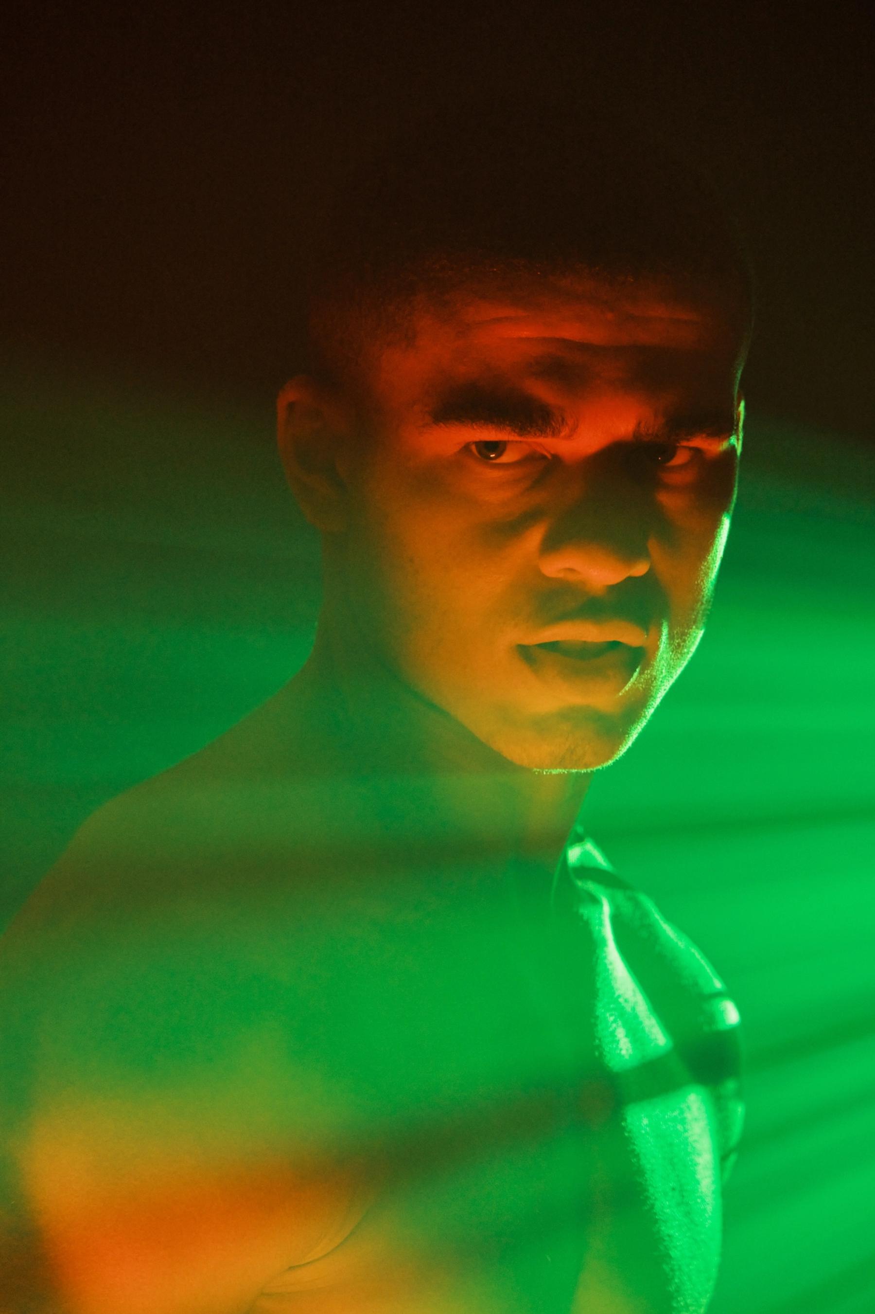 A man with a shirtless torso and dark hair stands in front of a green and red light, gazing directly at the camera.