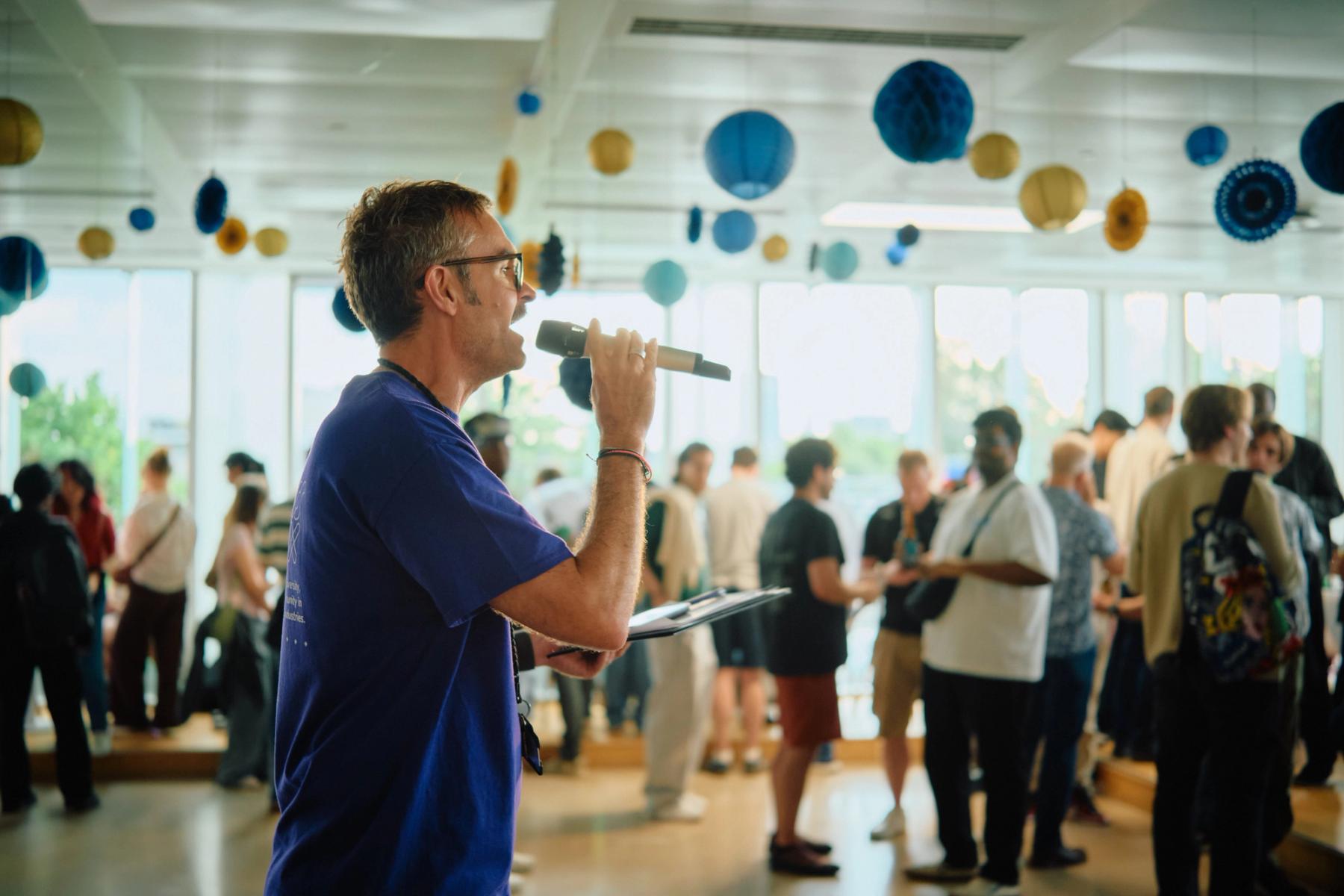Access VFX community event 2026, speaker in a blue shirt with clipboard addressing the room by large windows
